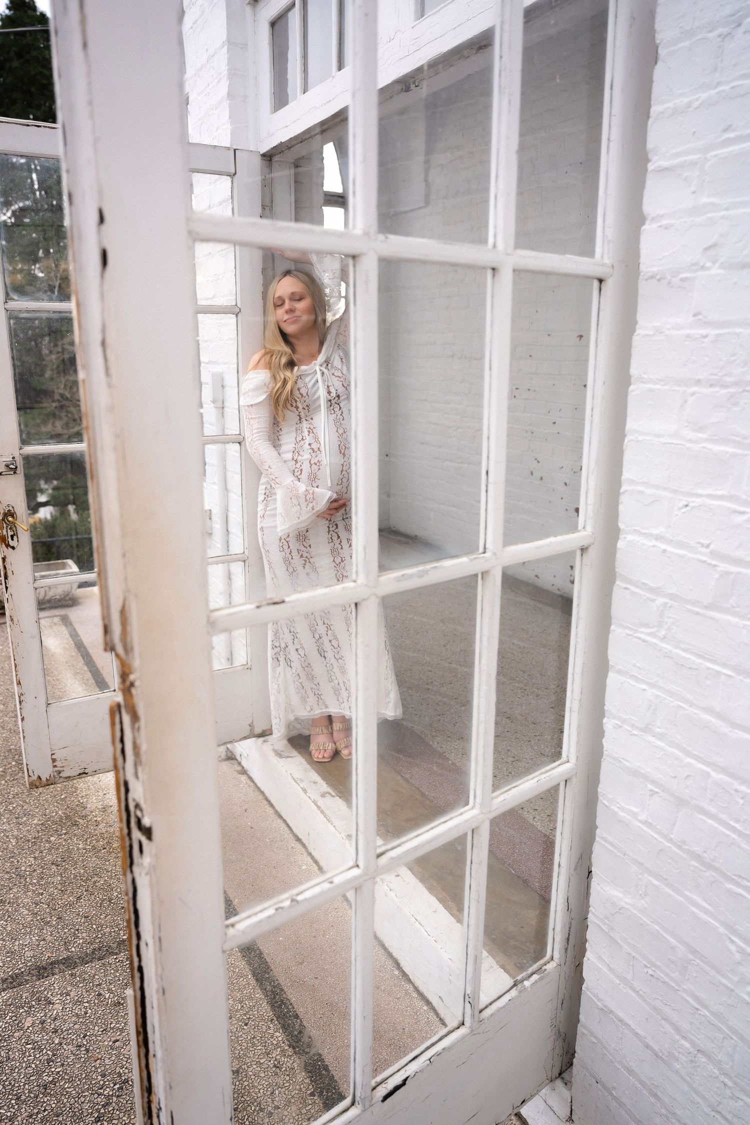 Woman in white lace dress poses by vintage white-framed windows.