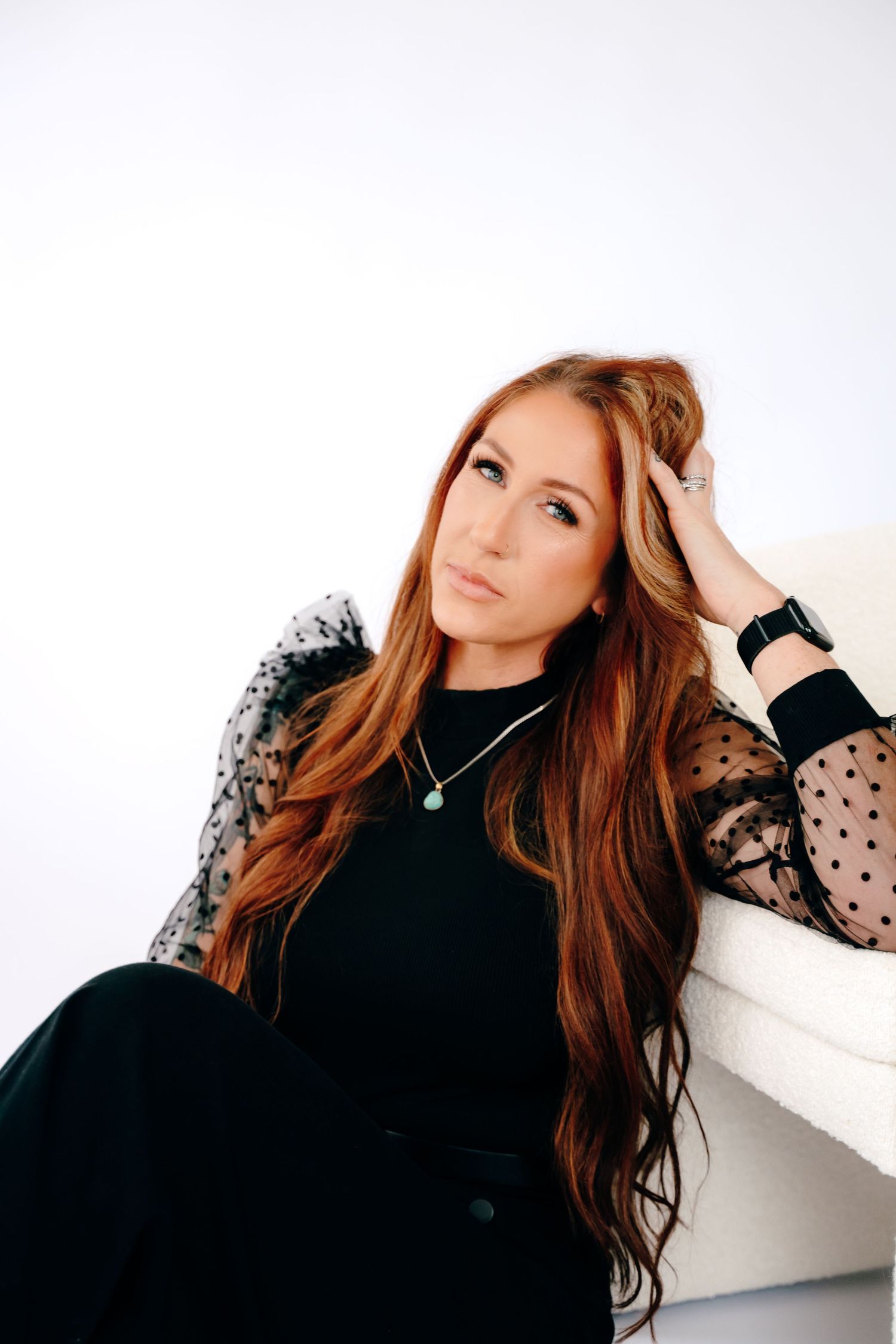 A woman with long auburn hair wearing a black dress with sheer polka dot sleeves poses against a white background.