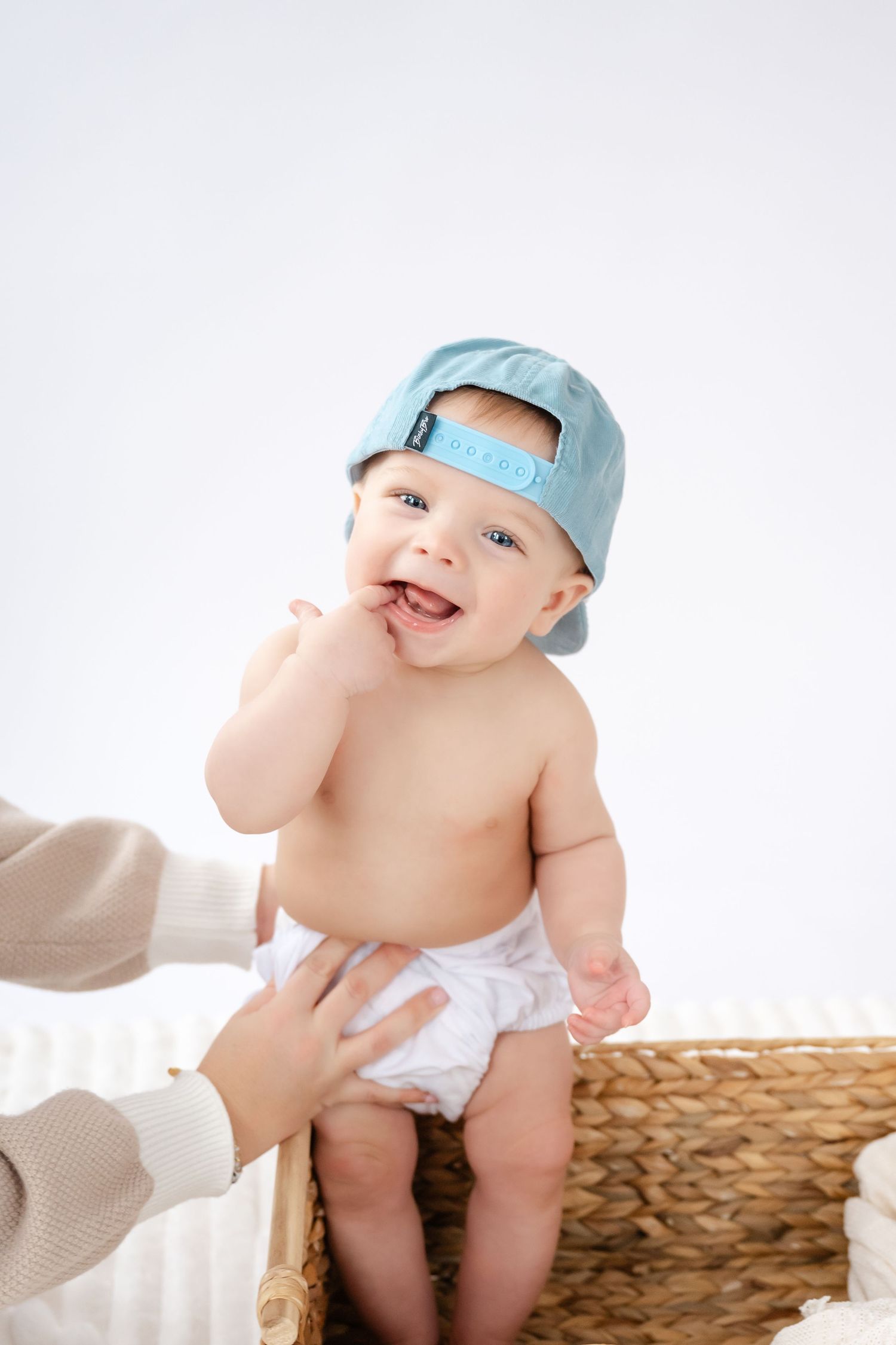Baby wearing light blue hat sits on woven basket while adult hands support them during photoshoot.