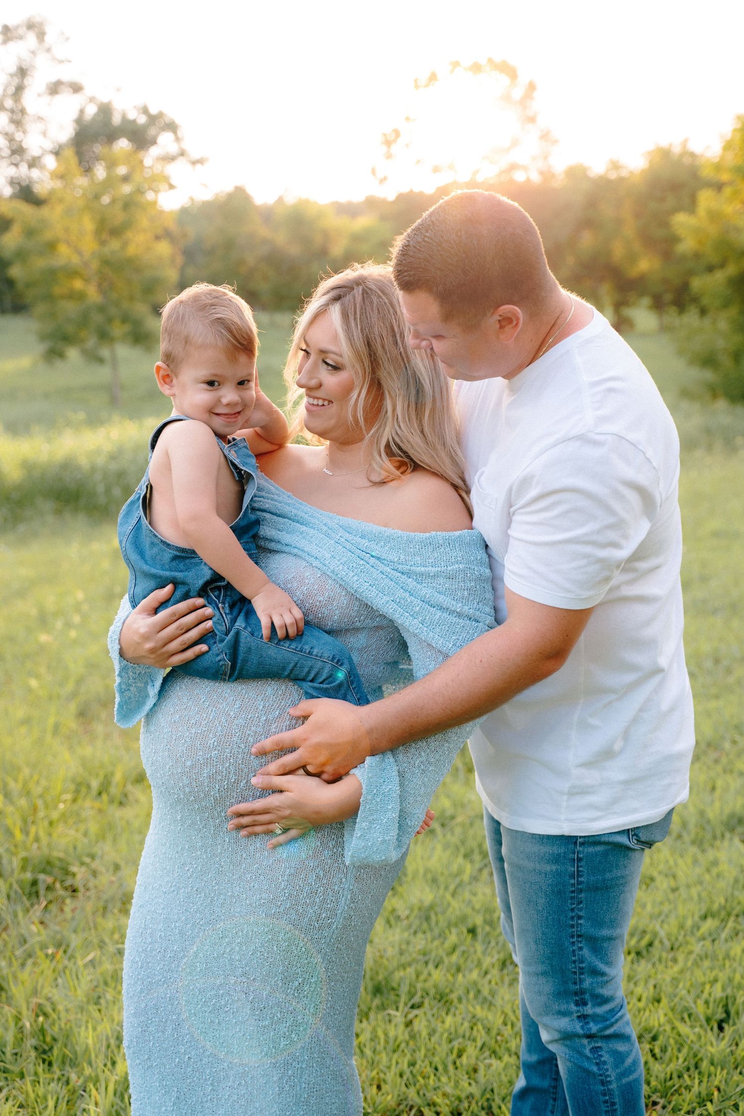 A family maternity session in a field at sunset with a toddler and parents.