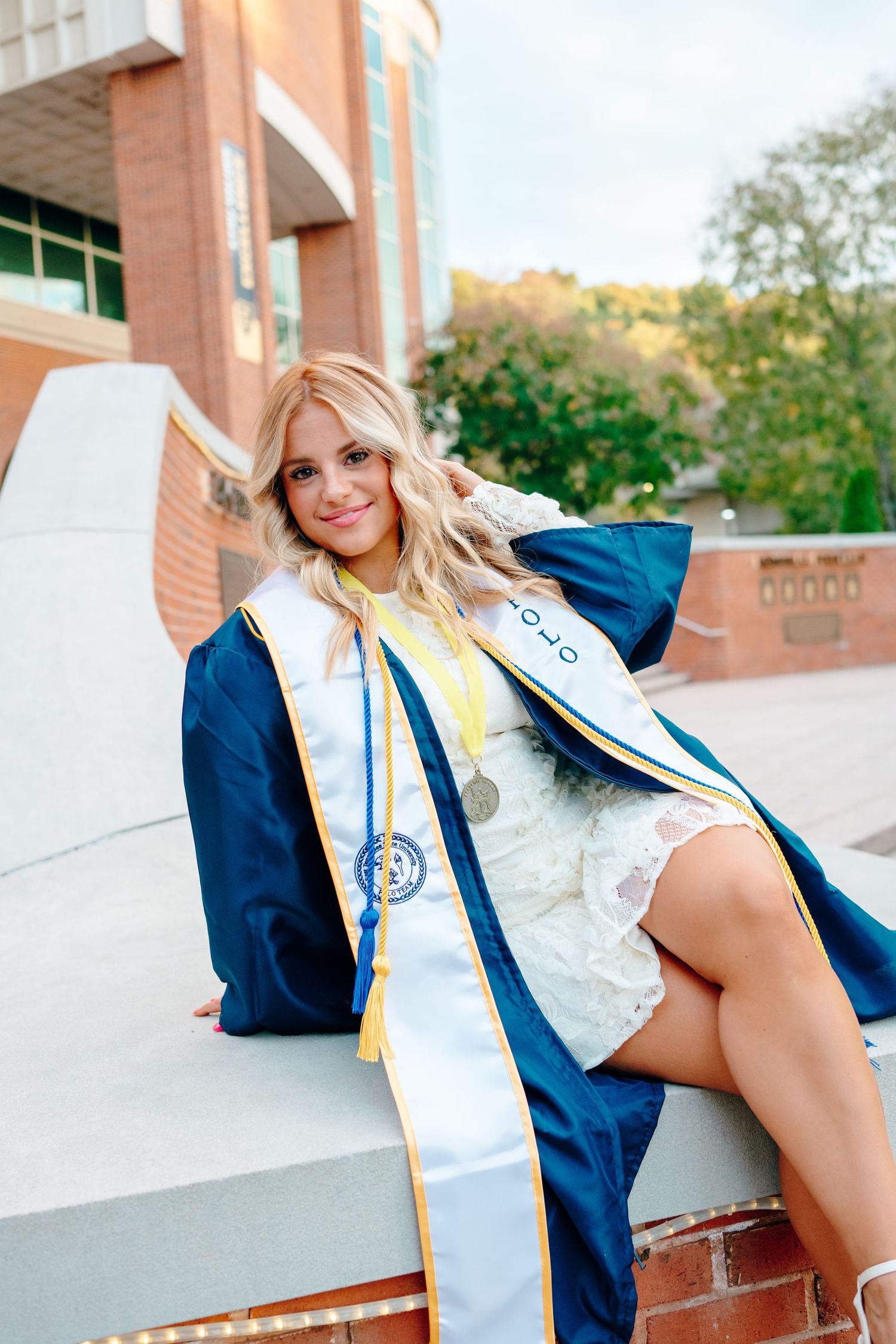 Graduate in white dress and blue graduation gown sits on concrete wall.