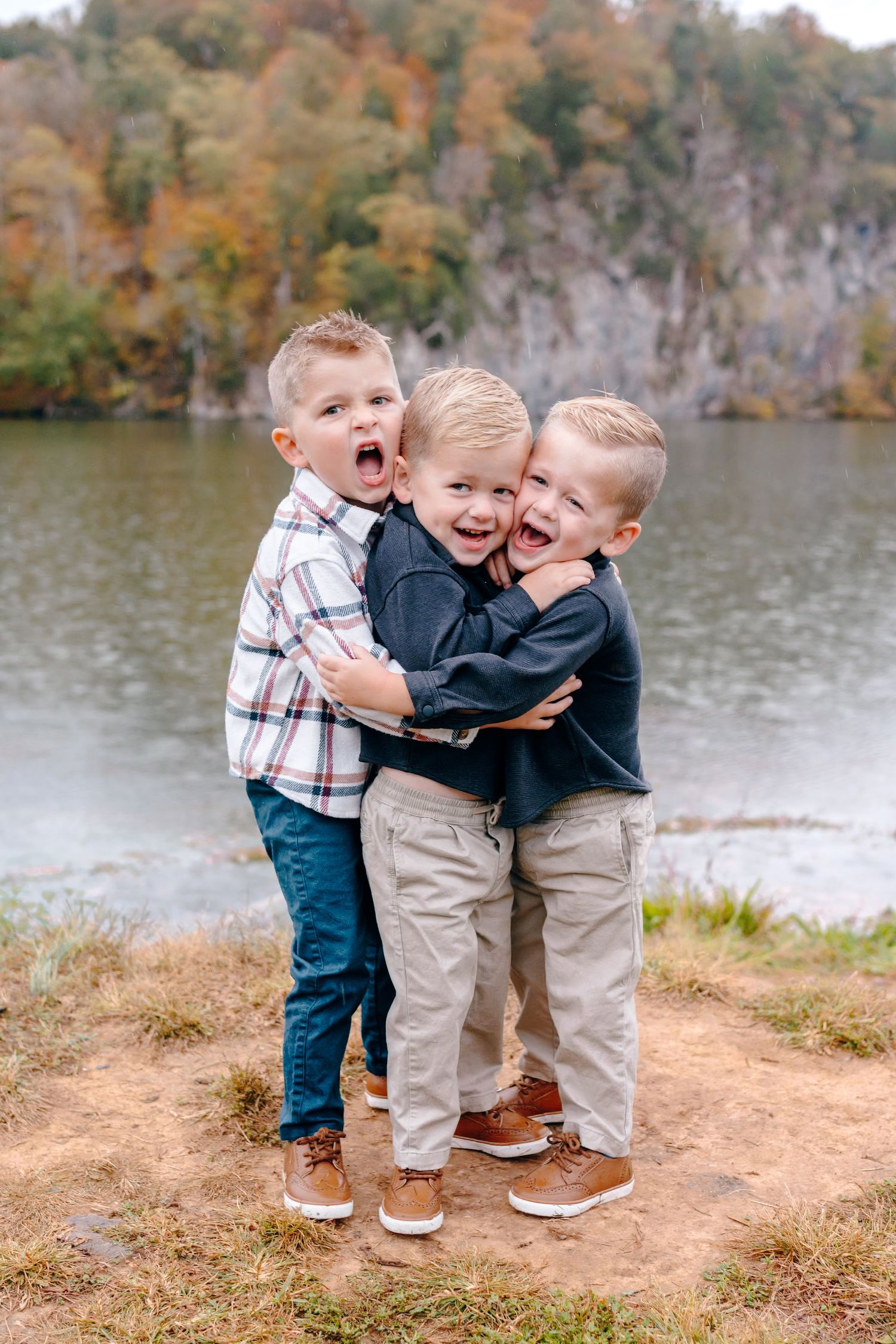 Three young children hugging and laughing by a lake with autumn foliage in background.