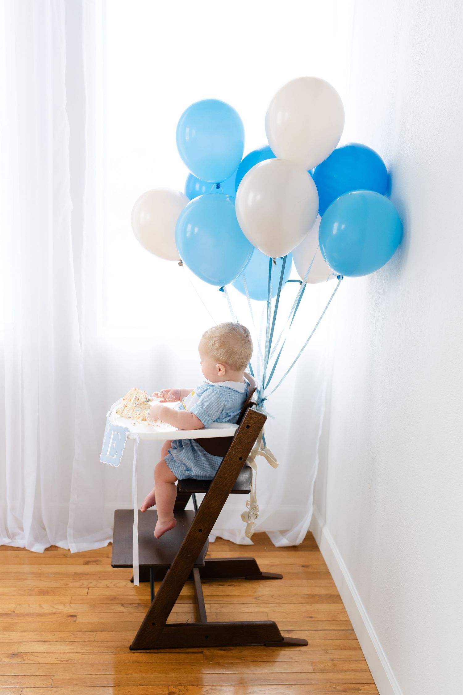 Baby sitting in high chair with blue and white balloons for first birthday celebration against white wall.