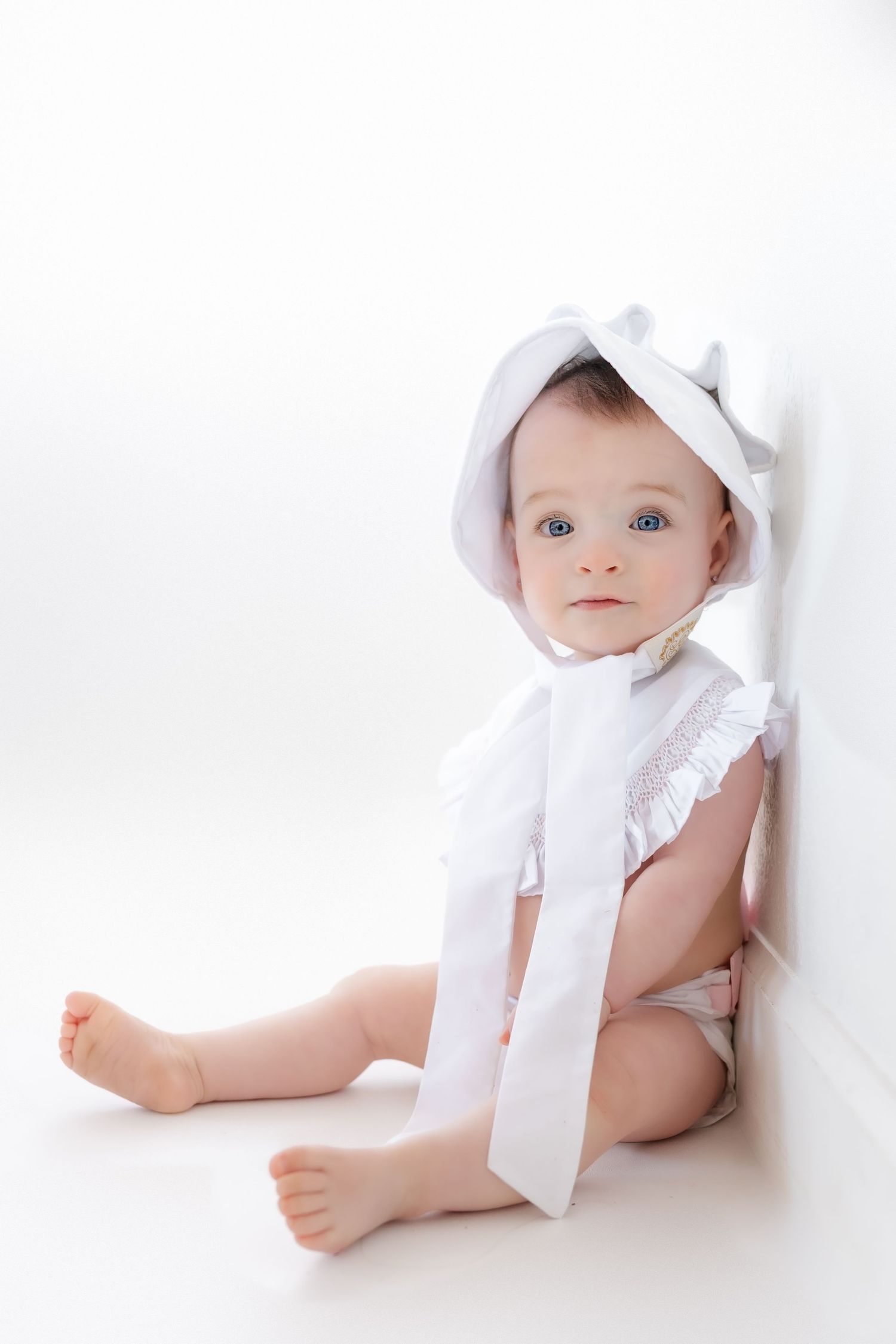 A baby wearing a white bonnet and dress lays on a white backdrop in a minimalist portrait series.