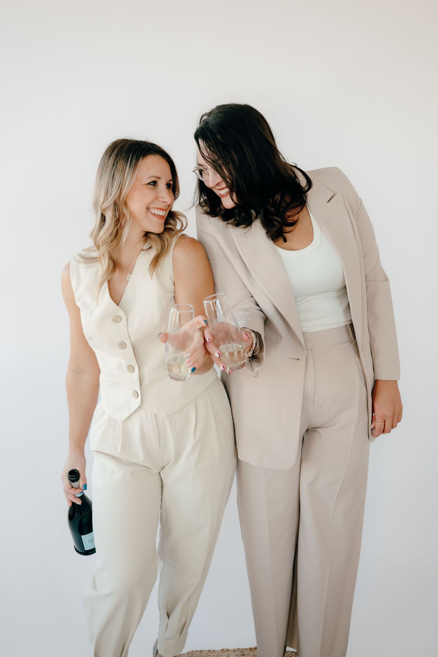 Two people in neutral-toned outfits share candid laughing moments in a minimalist white studio setting.