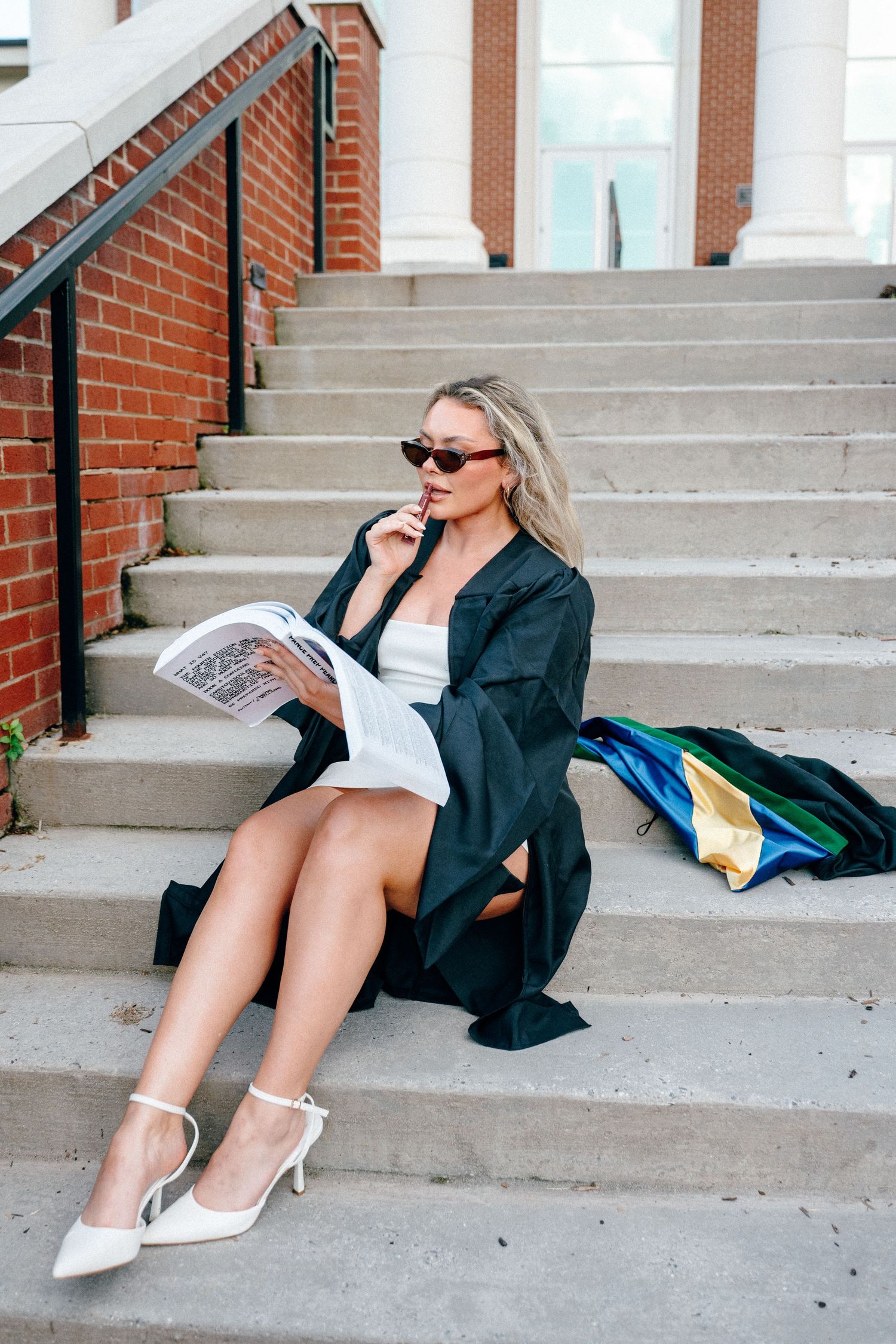 Graduate in black gown and sunglasses poses on concrete steps.