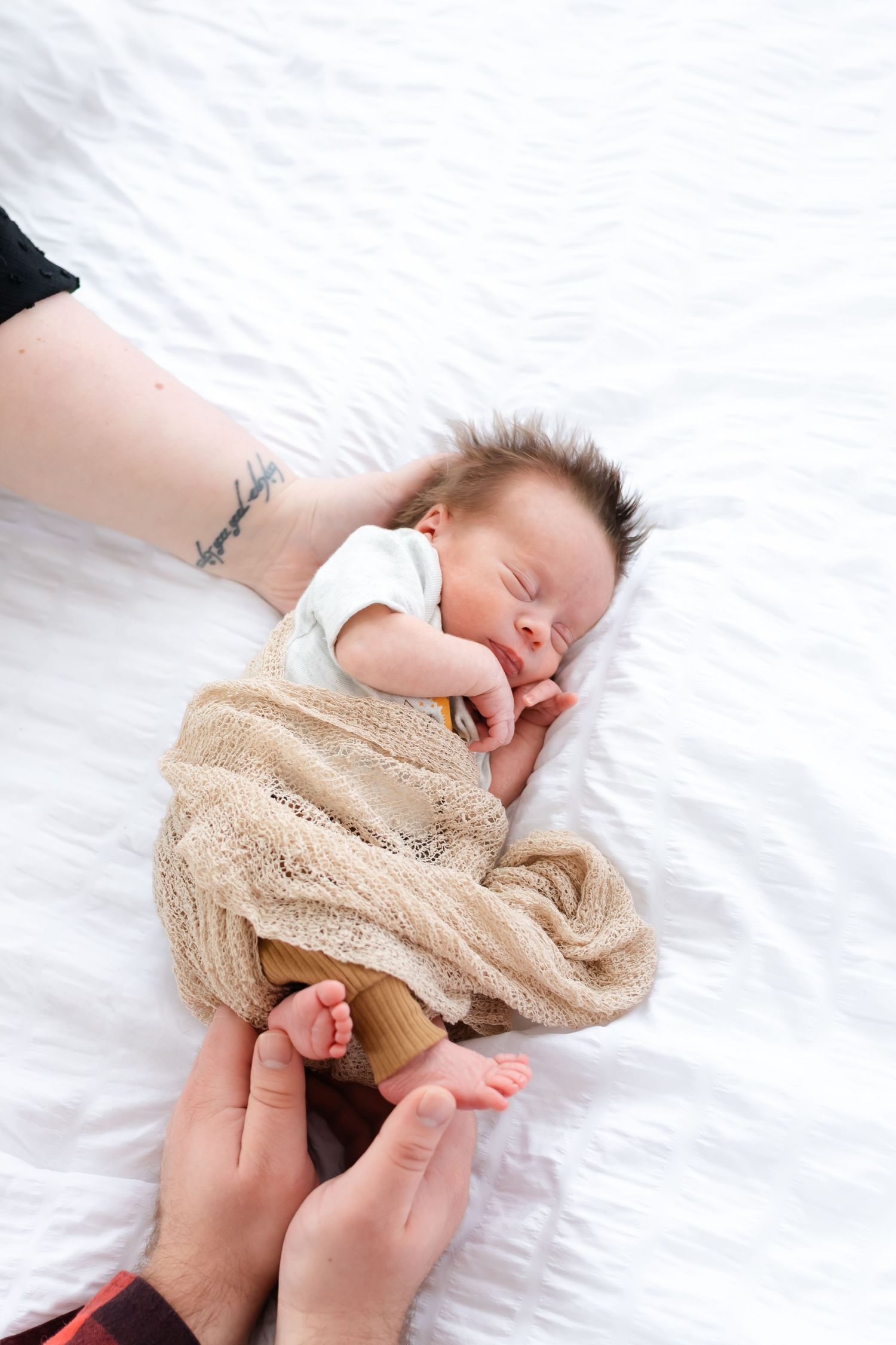 Baby wearing beige knit sweater lies on white bedding while being dressed.