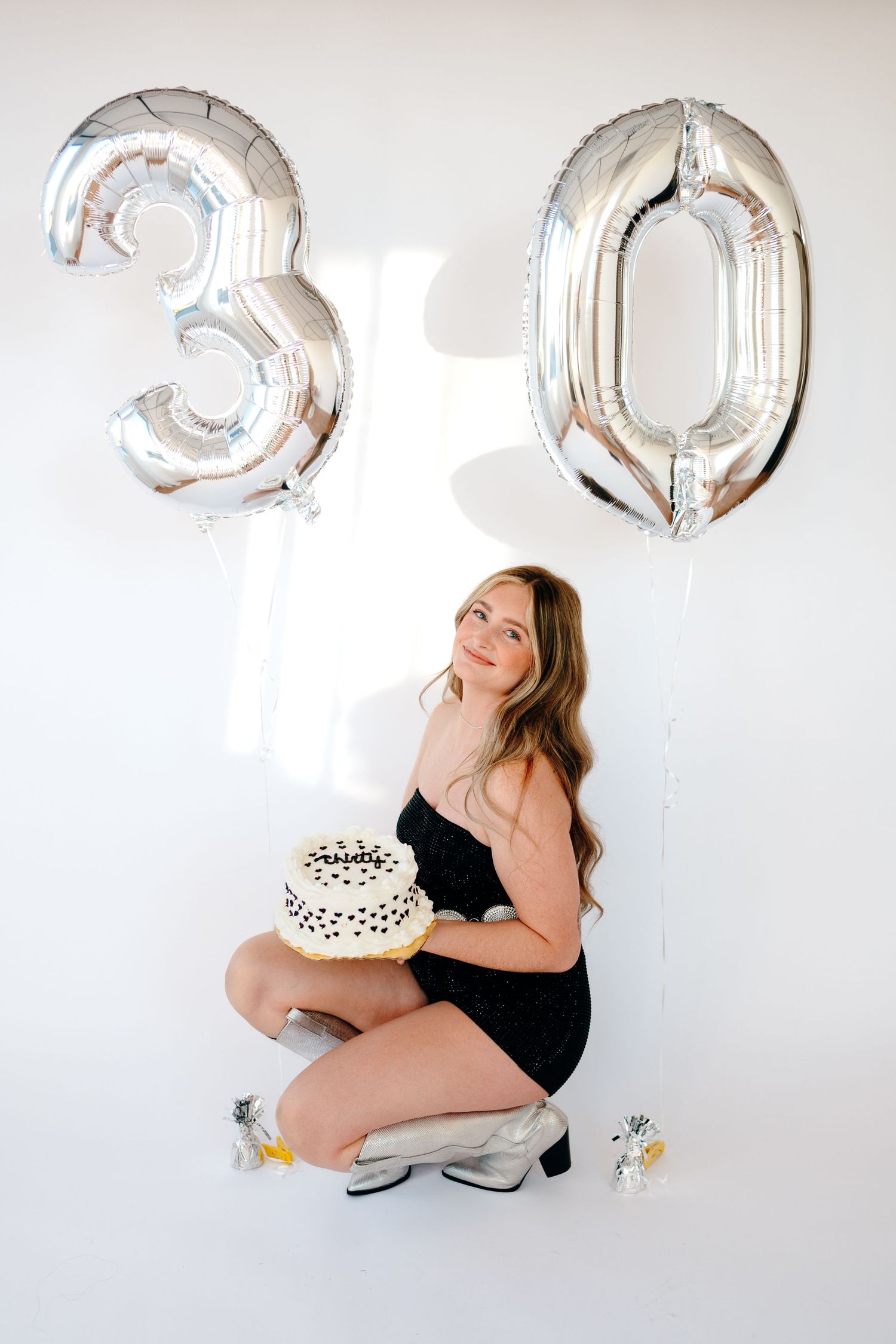 Maternity photoshoot with silver number balloons and white cake against light background.