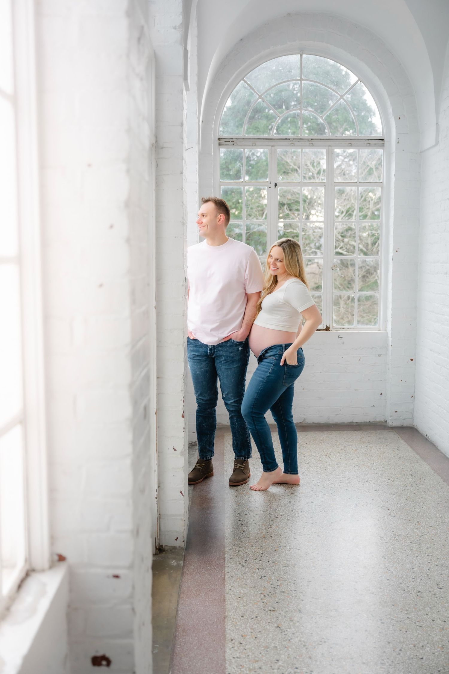 A couple playfully interacts in a bright white room with arched windows.