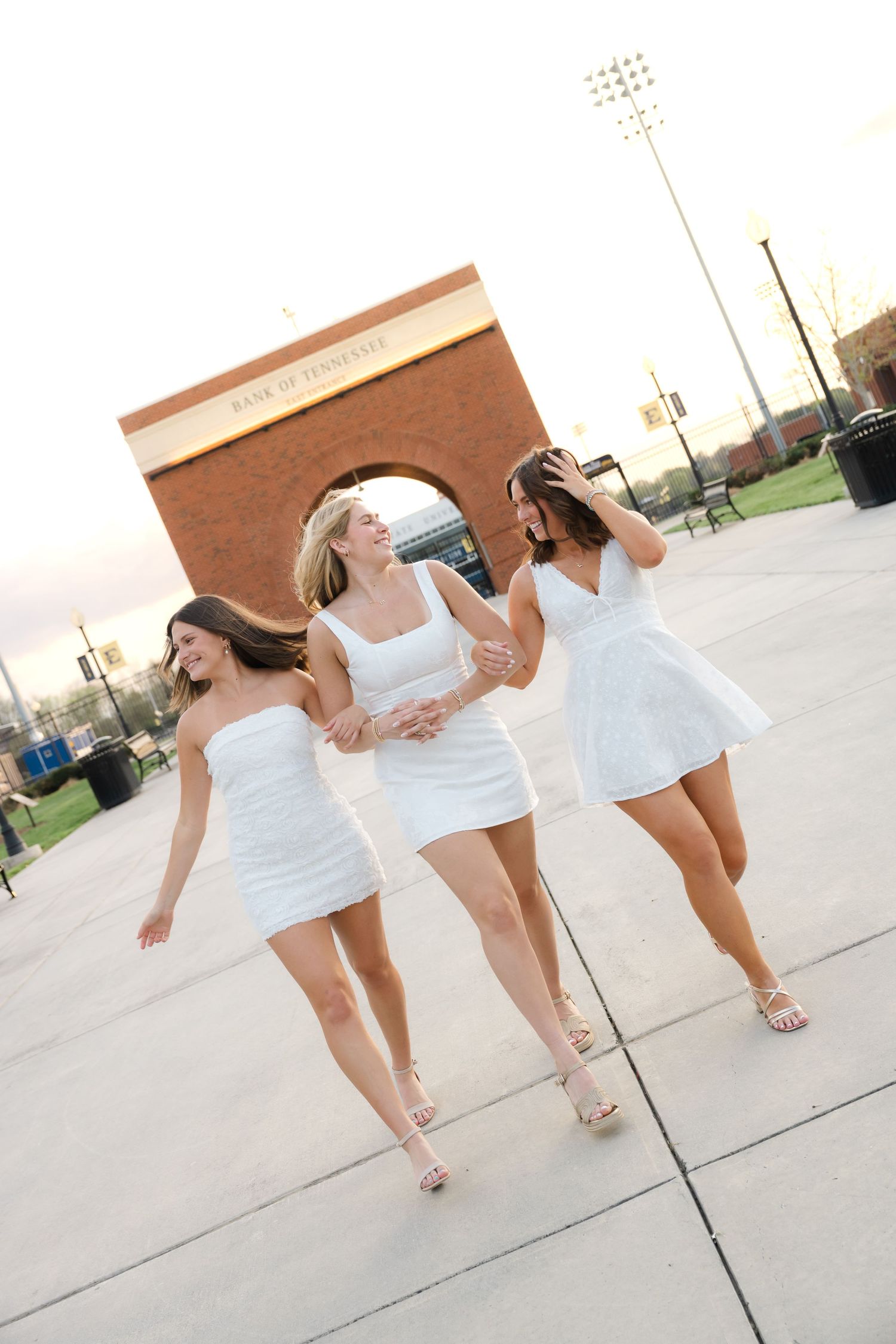 Three young women in white dresses walk together outdoors near a brick arch structure on a paved plaza.