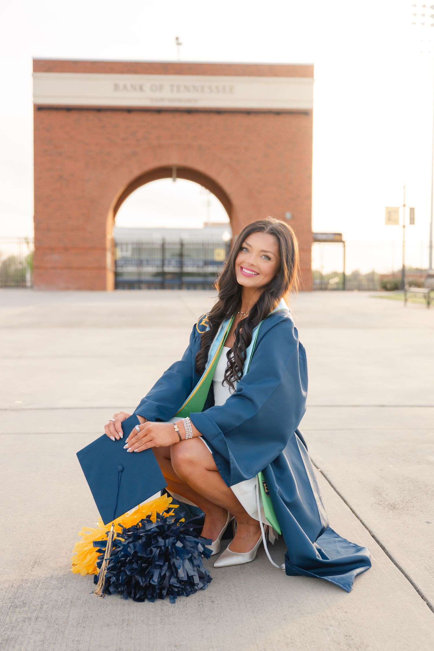 Young woman in blue graduation gown and cap smiling in front of Bank of Tennessee arch outdoors.