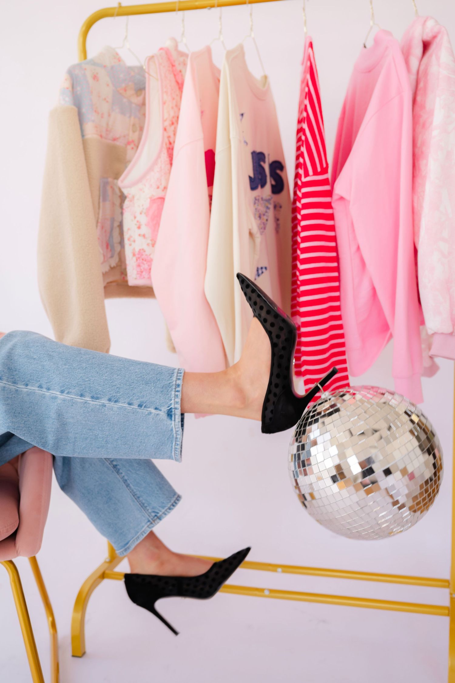 Legs wearing blue jeans and black heels pose against a pink clothing rack with a disco ball in studio setting.