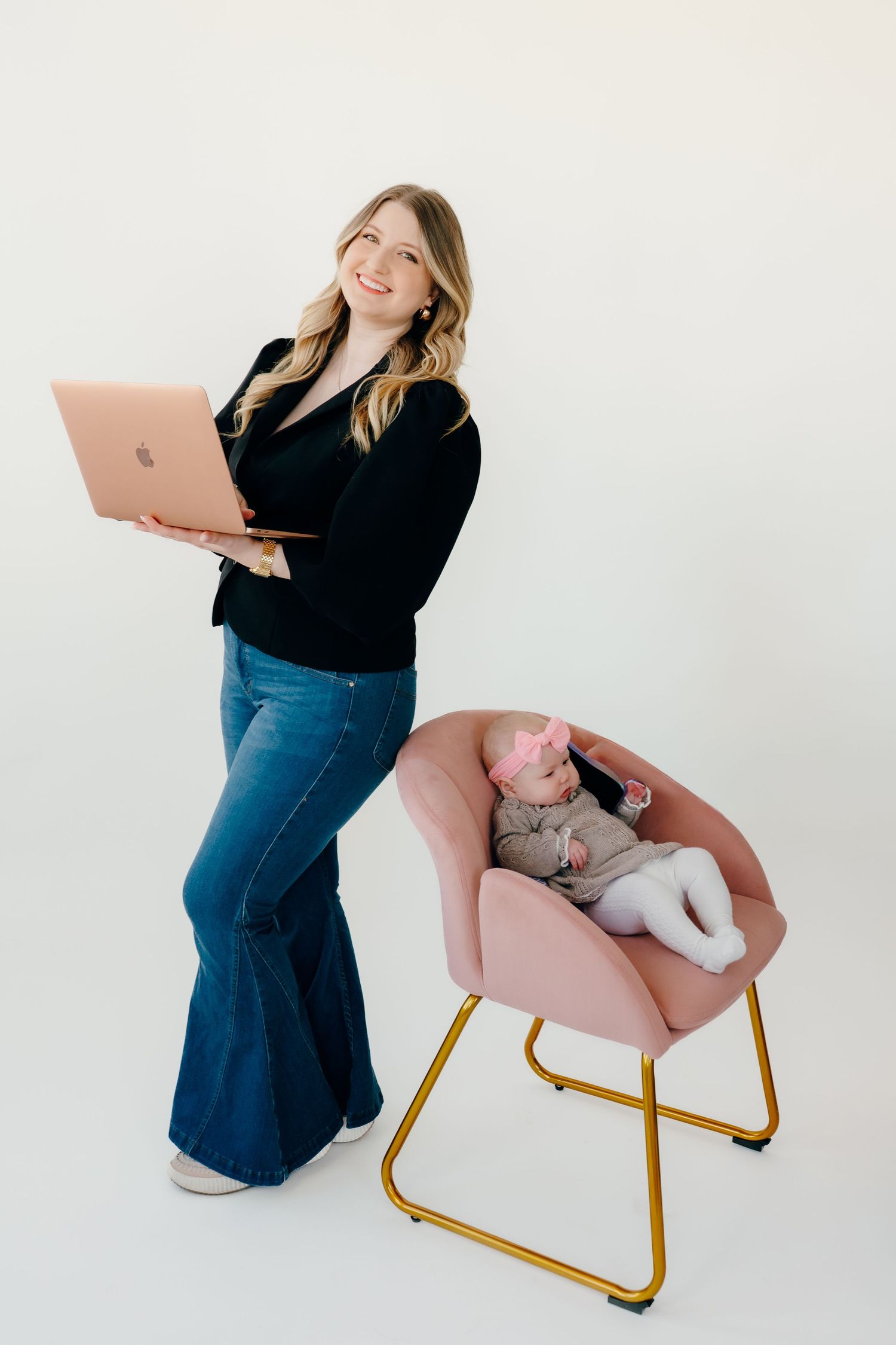 Person in black sweater and jeans holds laptop while standing next to pink chair with sleeping cat.