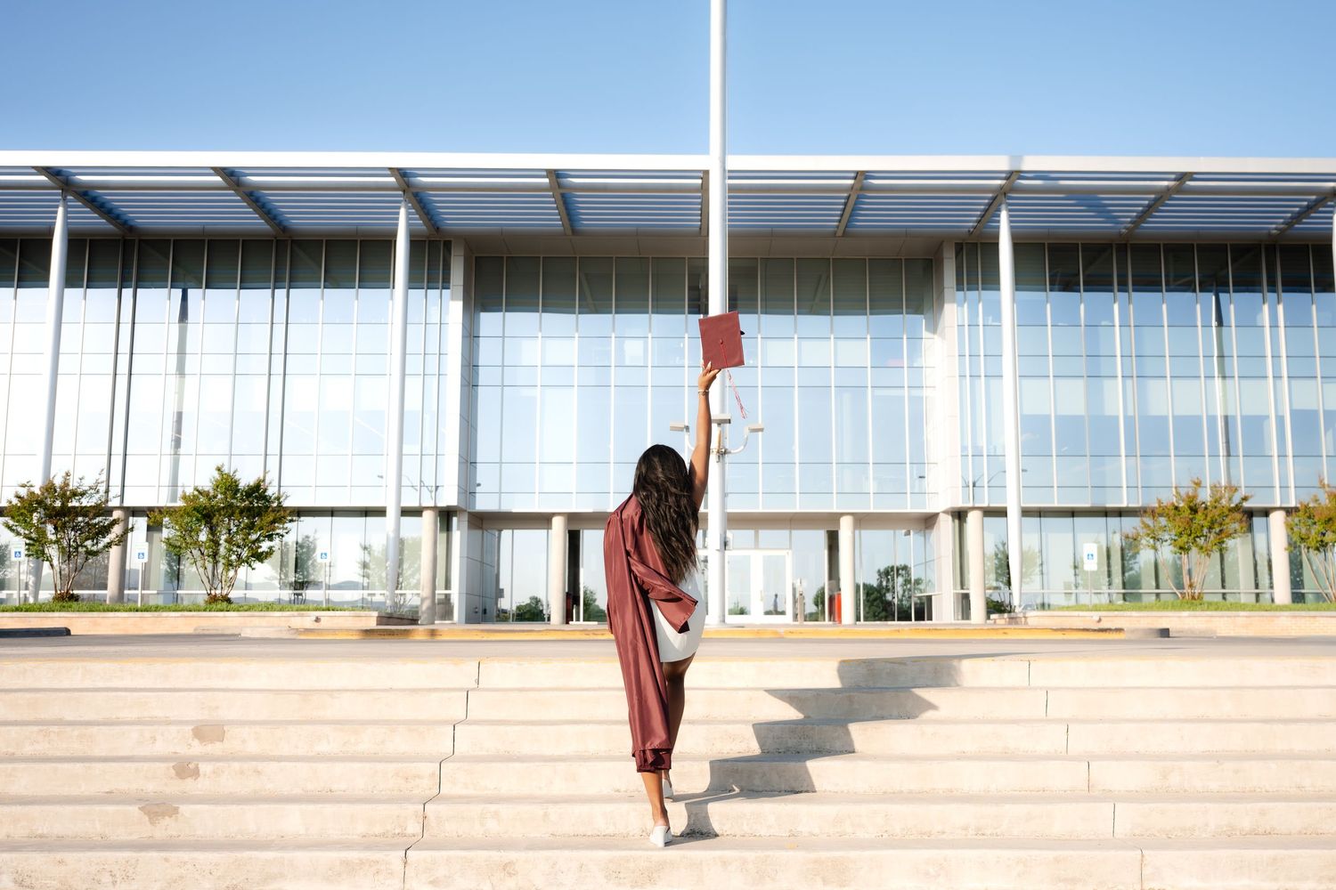 Graduate in red gown raises diploma on university steps, celebrating academic achievement outdoors.