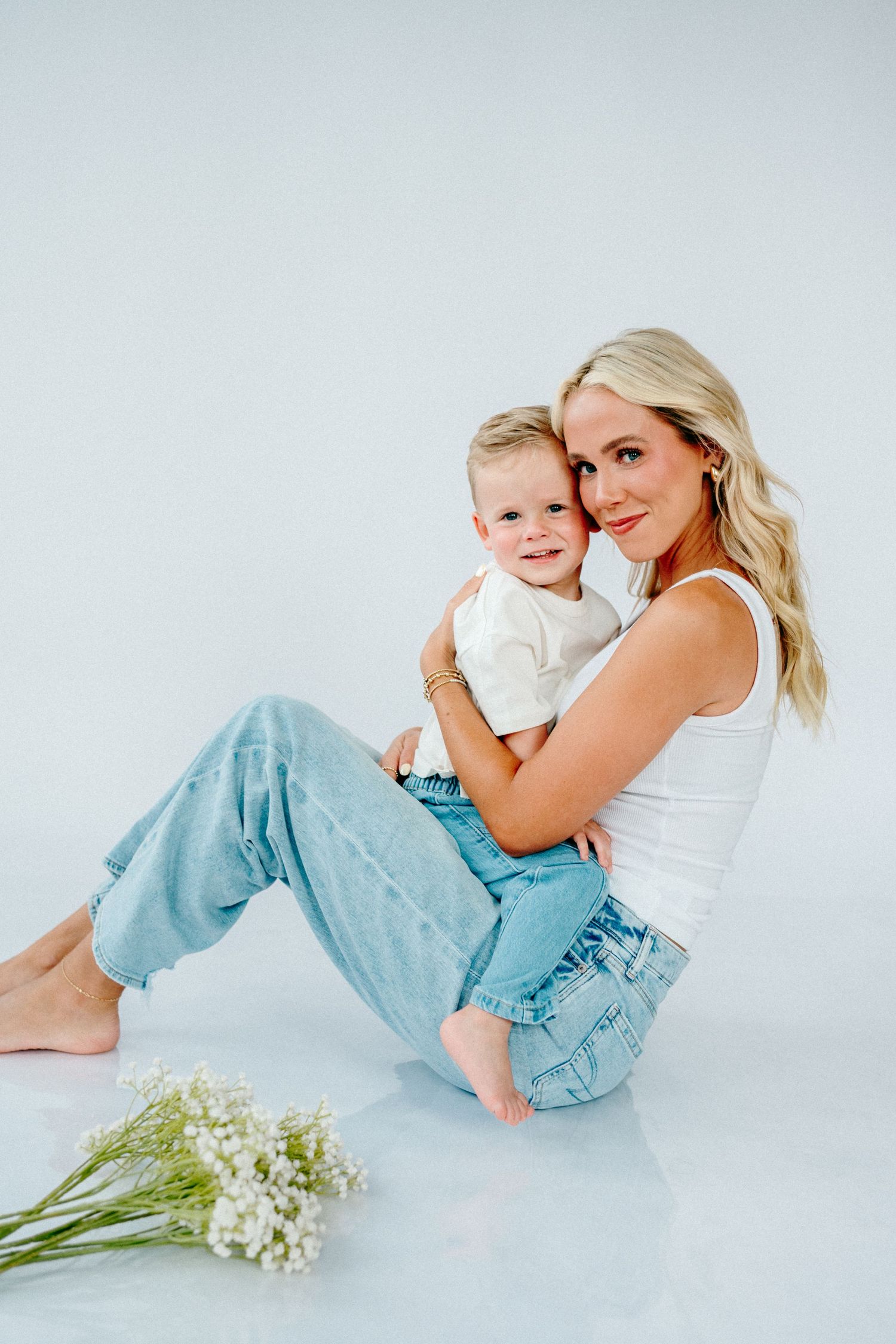Mother and child in white and blue outfits pose playfully together on white background with lily of the valley flowers.