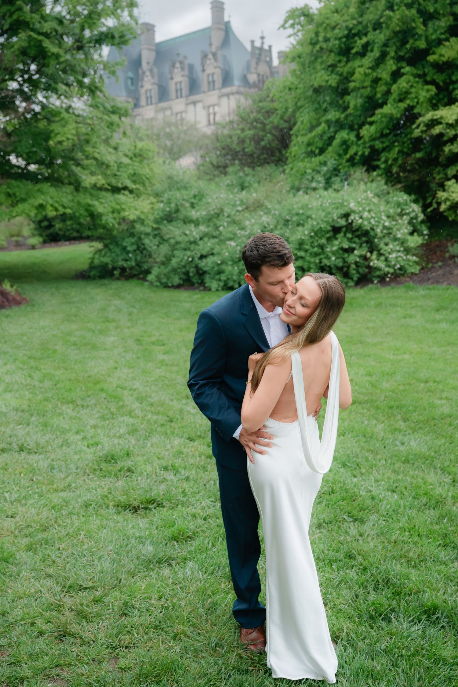 A couple shares a romantic embrace on green lawn with a grand estate building and lush trees in the background.
