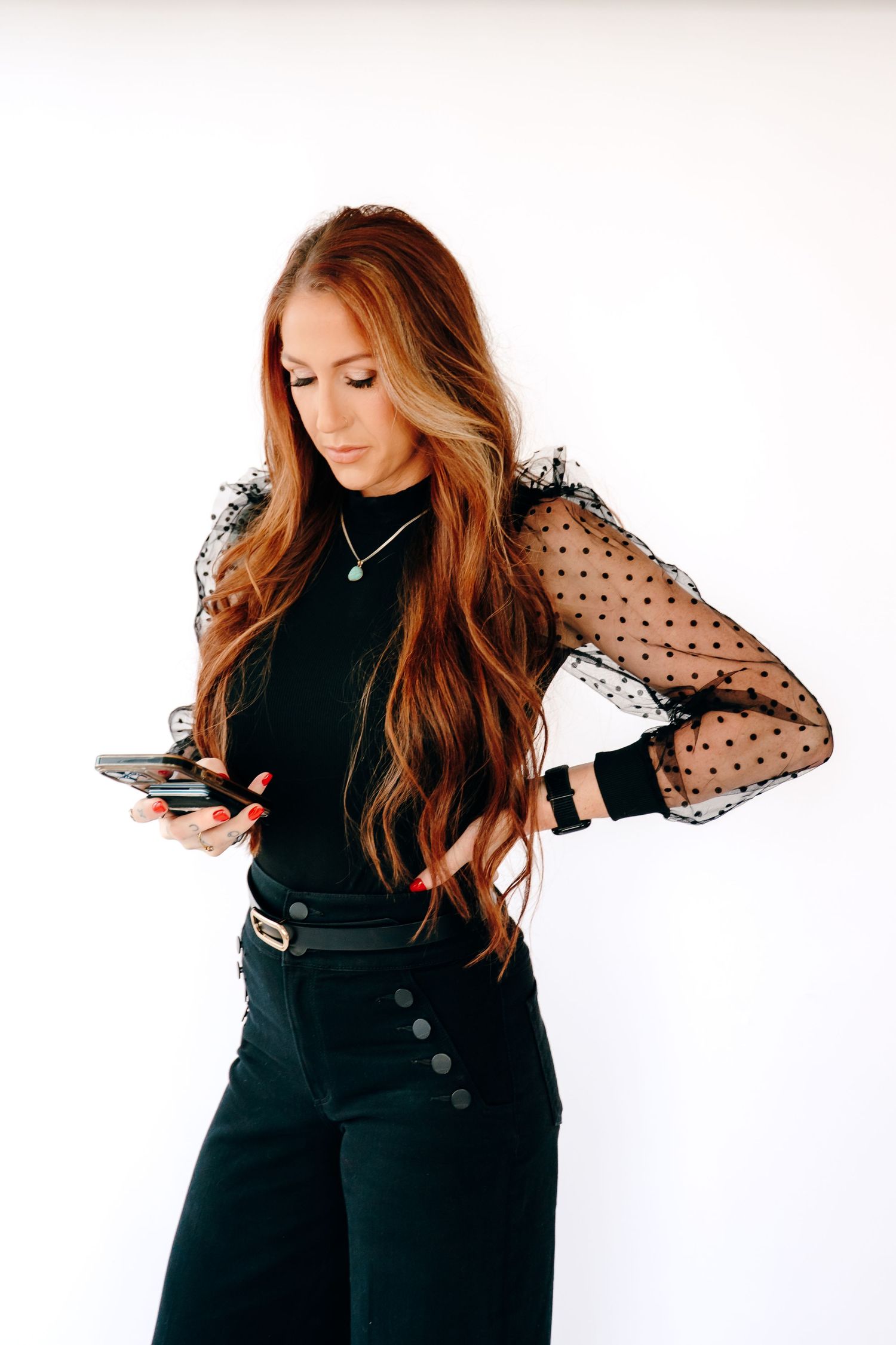Stylish black outfit featuring sheer polka dot sleeves and high waisted pants while checking phone against white wall.