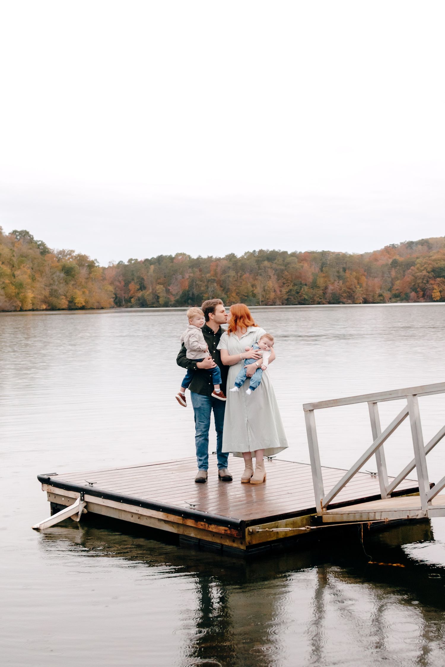 Family of four sharing a kiss on a wooden dock by a lake surrounded by autumn foliage on an overcast day.