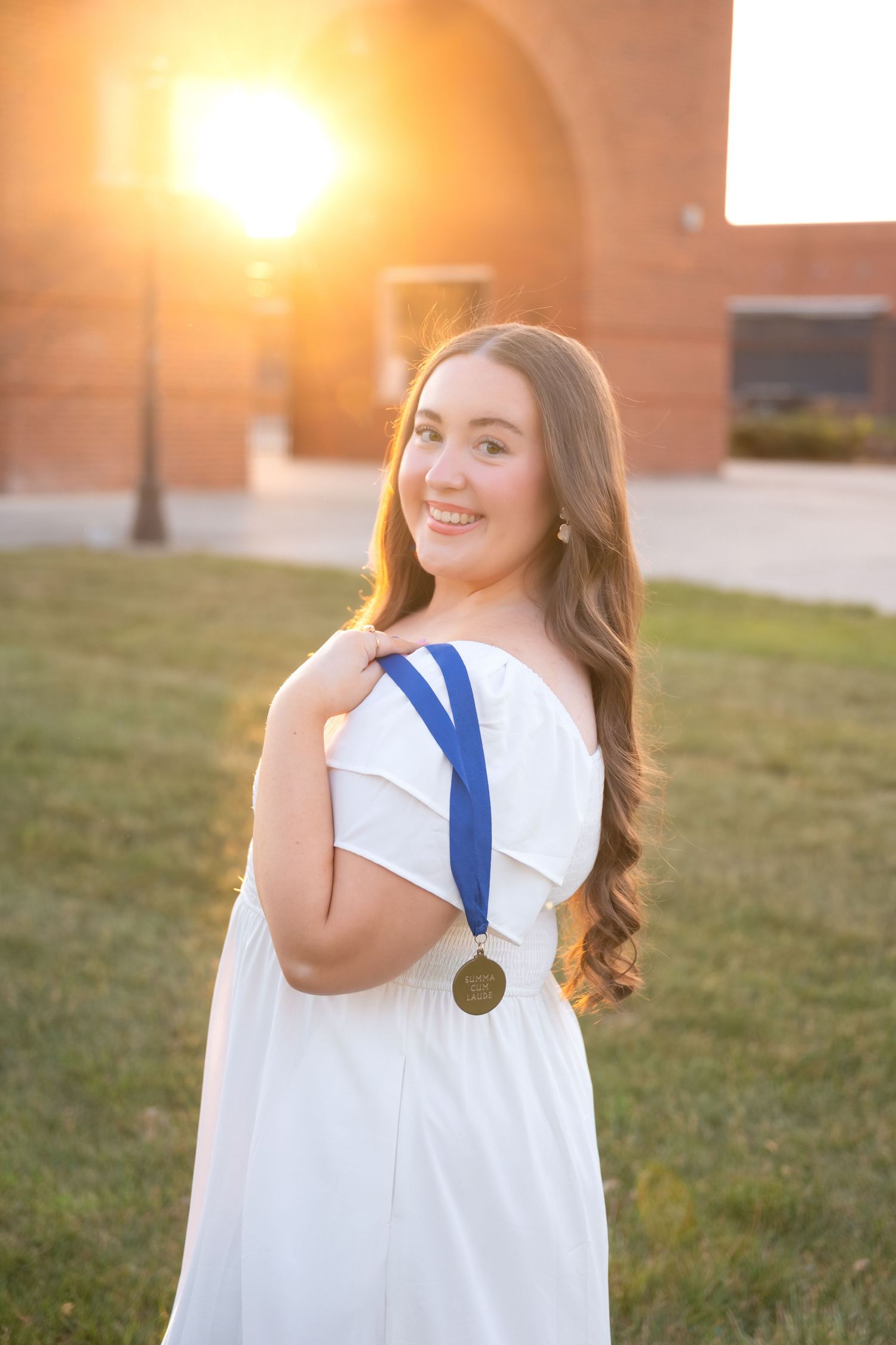 Young woman in white off-shoulder dress displaying gold medal with blue ribbon, posing outdoors at golden hour sunset.