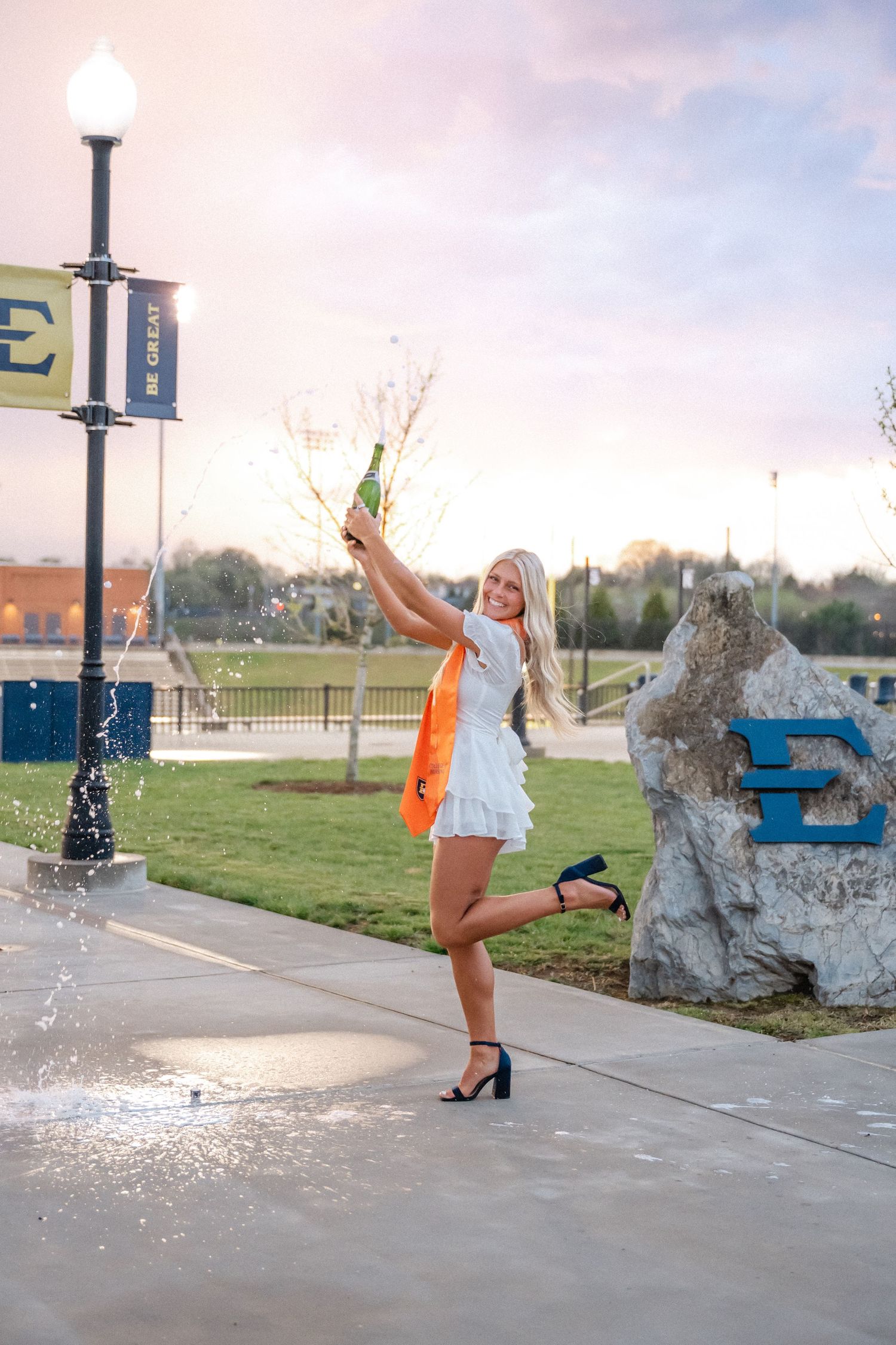 Excited blonde graduate in white dress popping champagne bottle near ETSU campus sign at sunset.