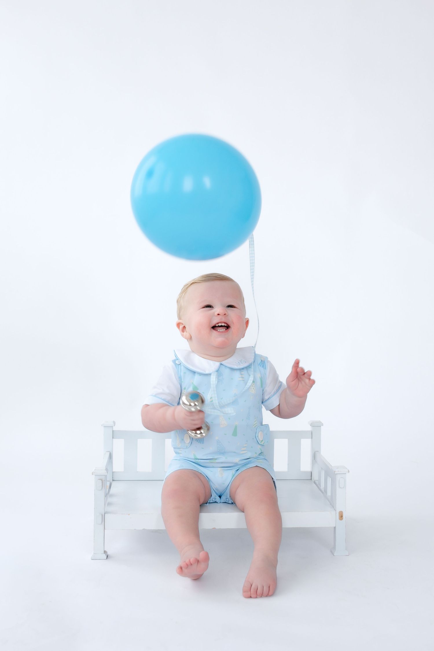 Baby in light blue outfit sits on white bench with blue balloon playfully covering their face.