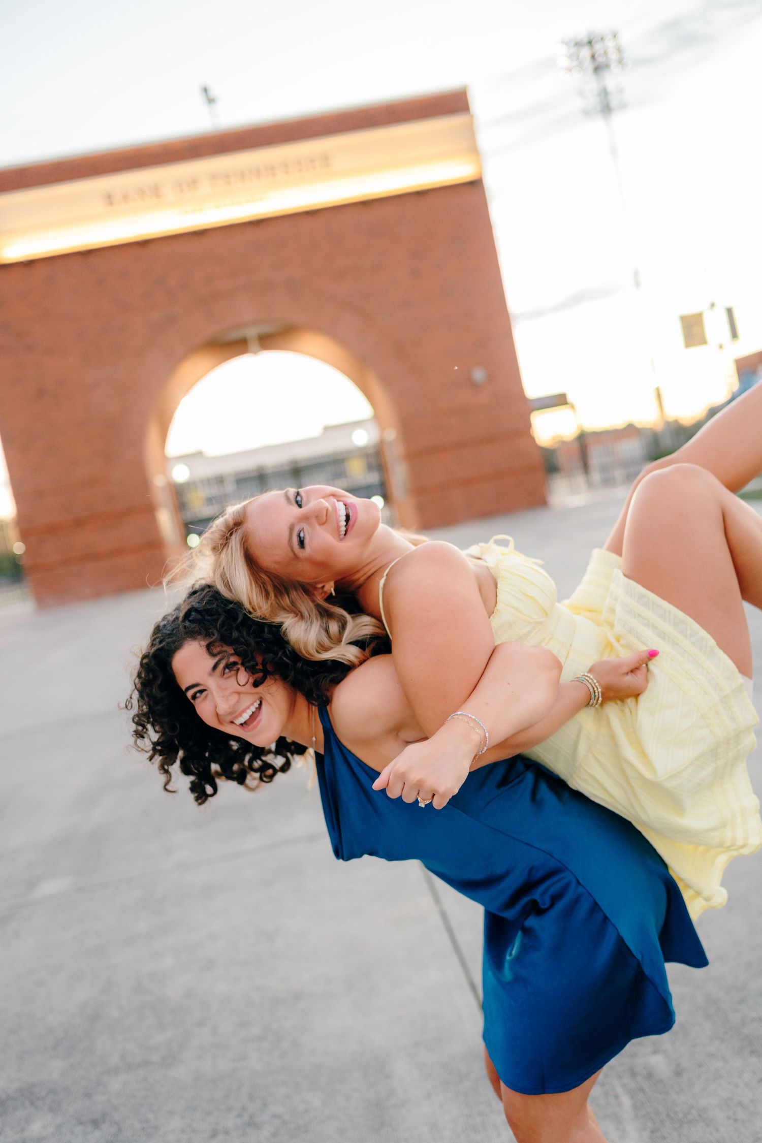 Two friends in yellow and blue dresses playfully pose in front of a brick archway at sunset.