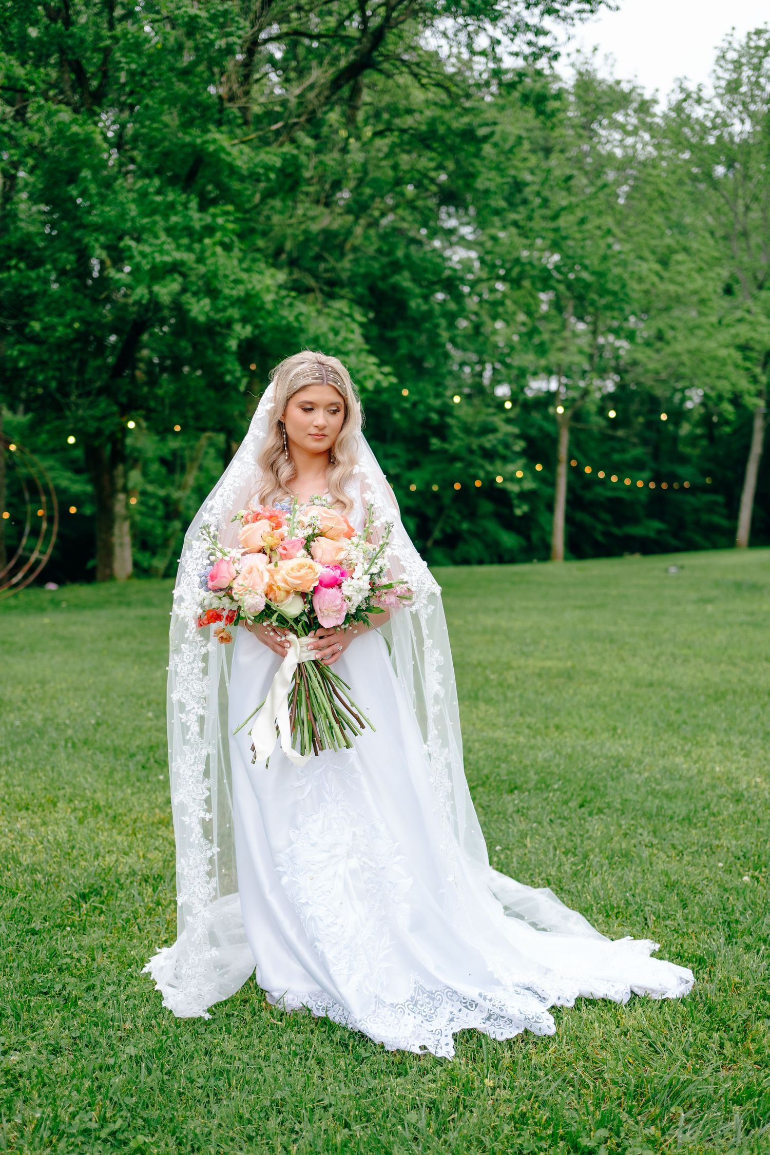 Bride in white lace dress holds vibrant pink and coral bouquet on green lawn.