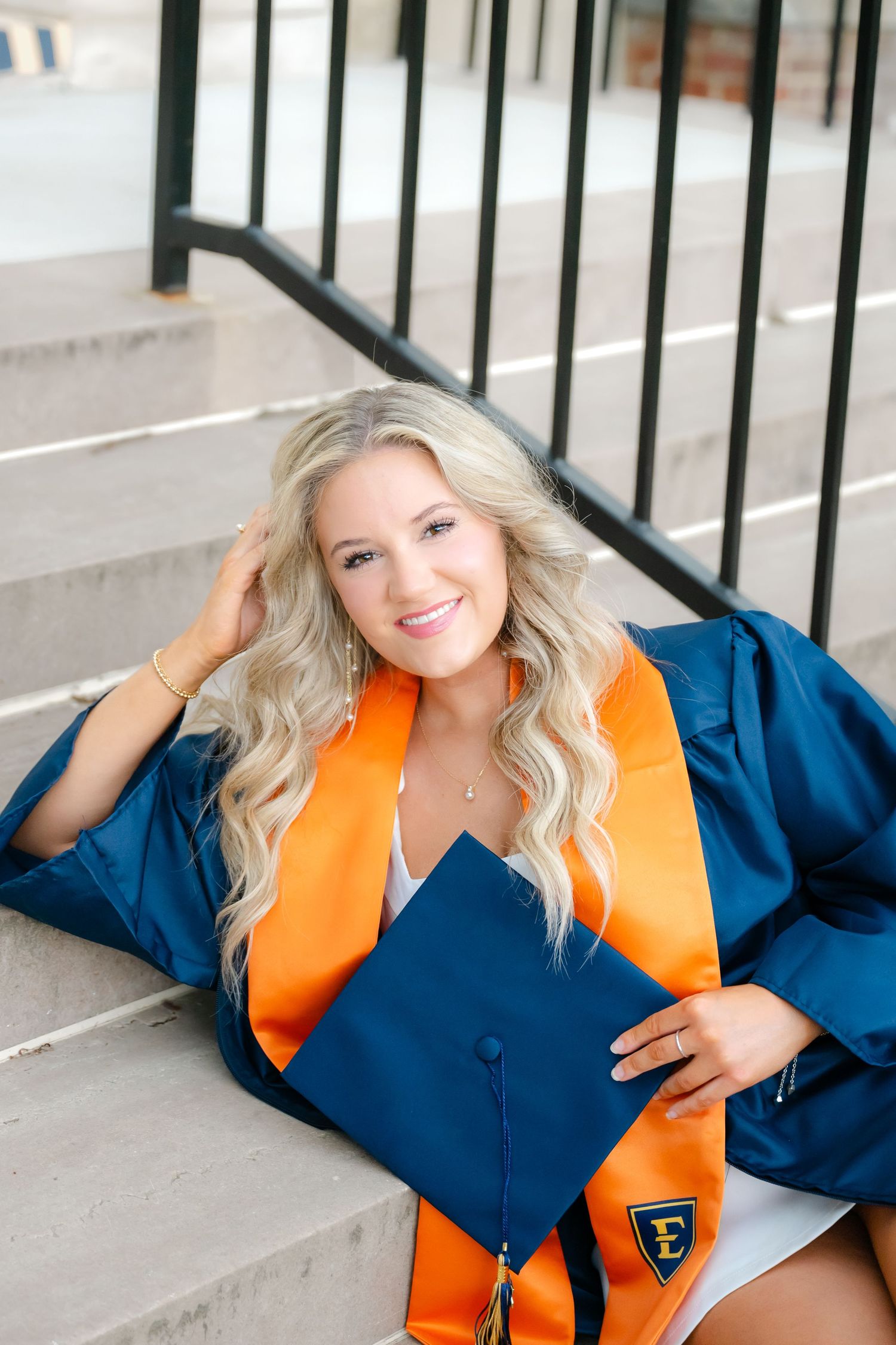 Smiling blonde woman in blue graduation gown and orange sash sits on steps holding cap.