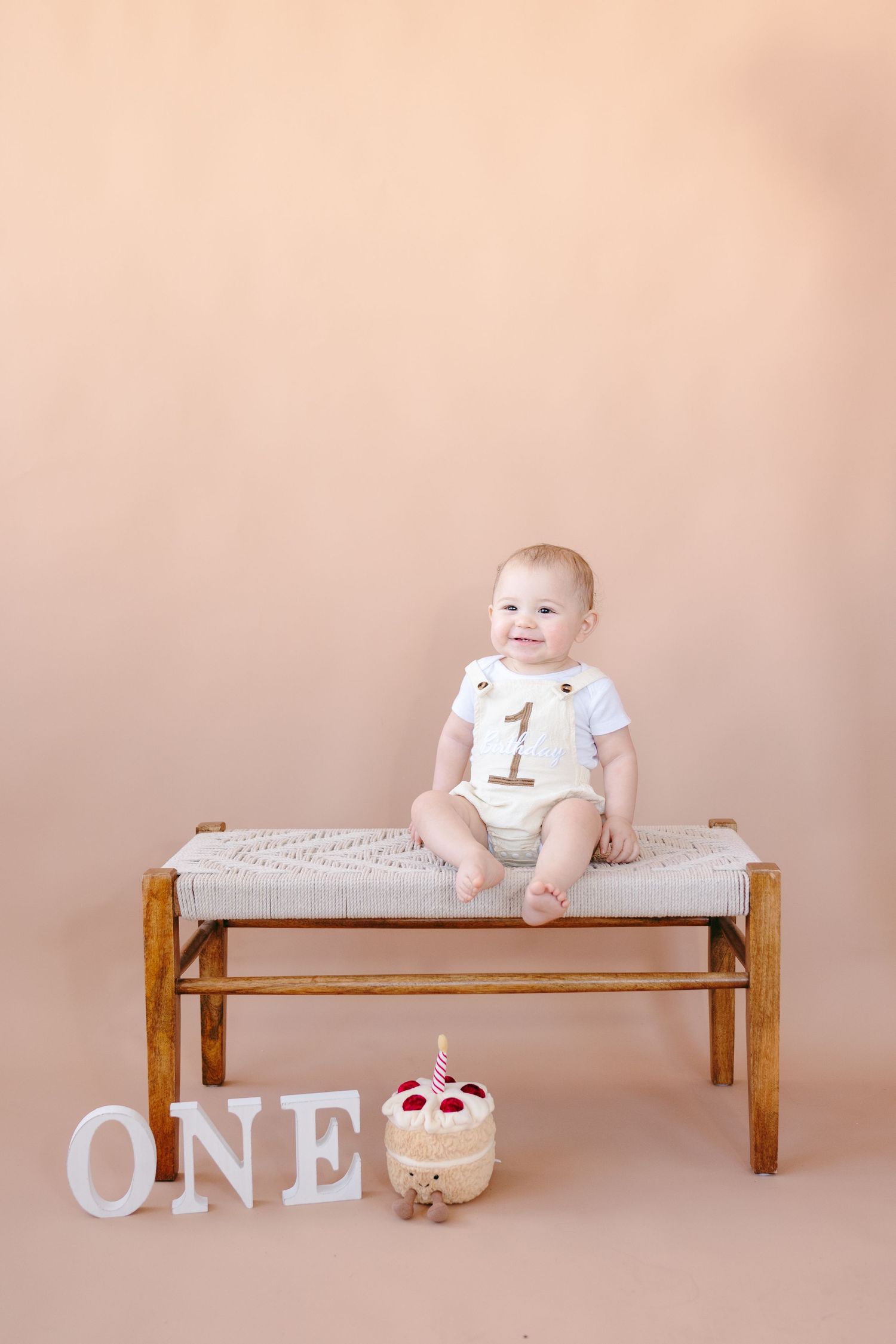 Baby sits on wooden bench with 'ONE' letters and decorative cake display.
