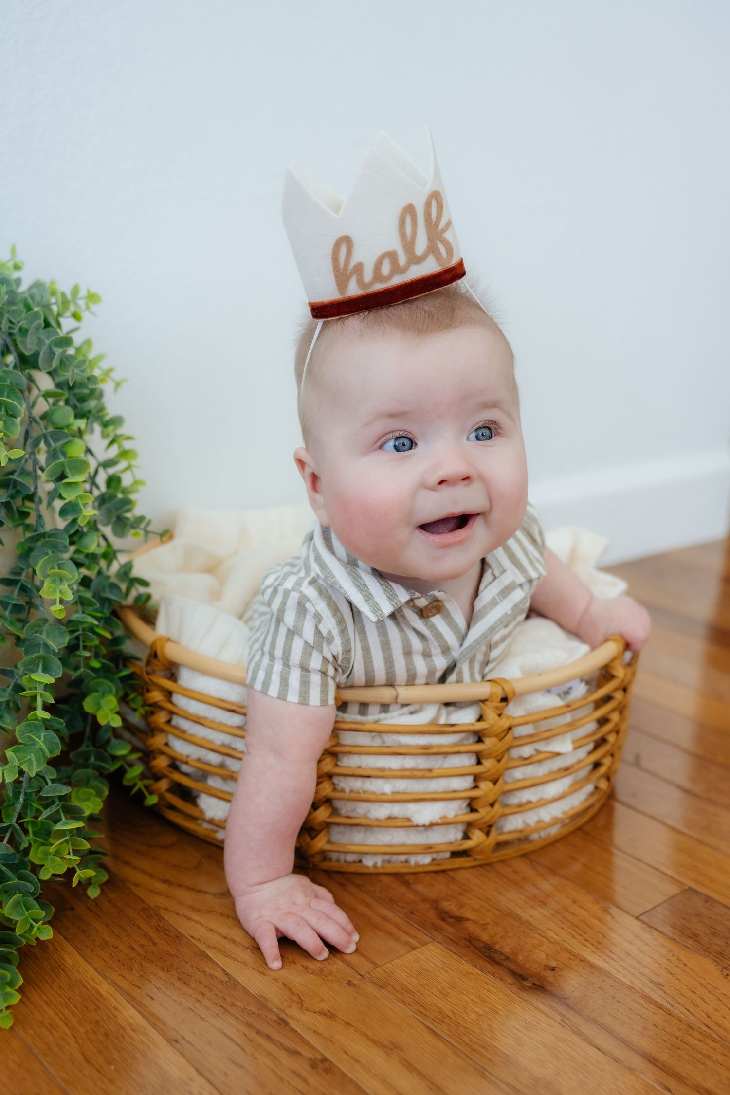Baby in striped outfit and paper crown sitting in woven basket on hardwood floor.