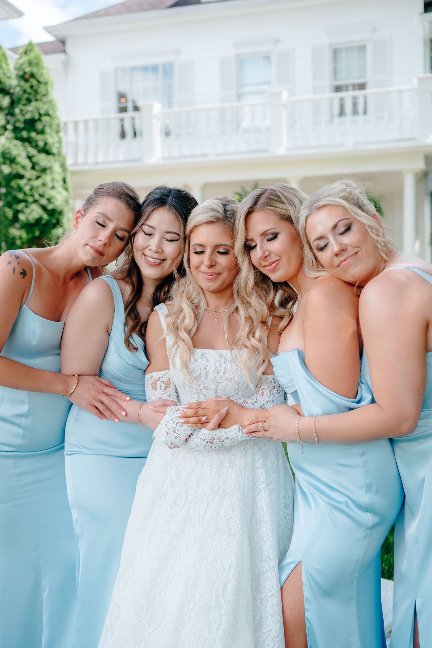 Bridal party in matching light blue dresses embraces on mansion lawn.
