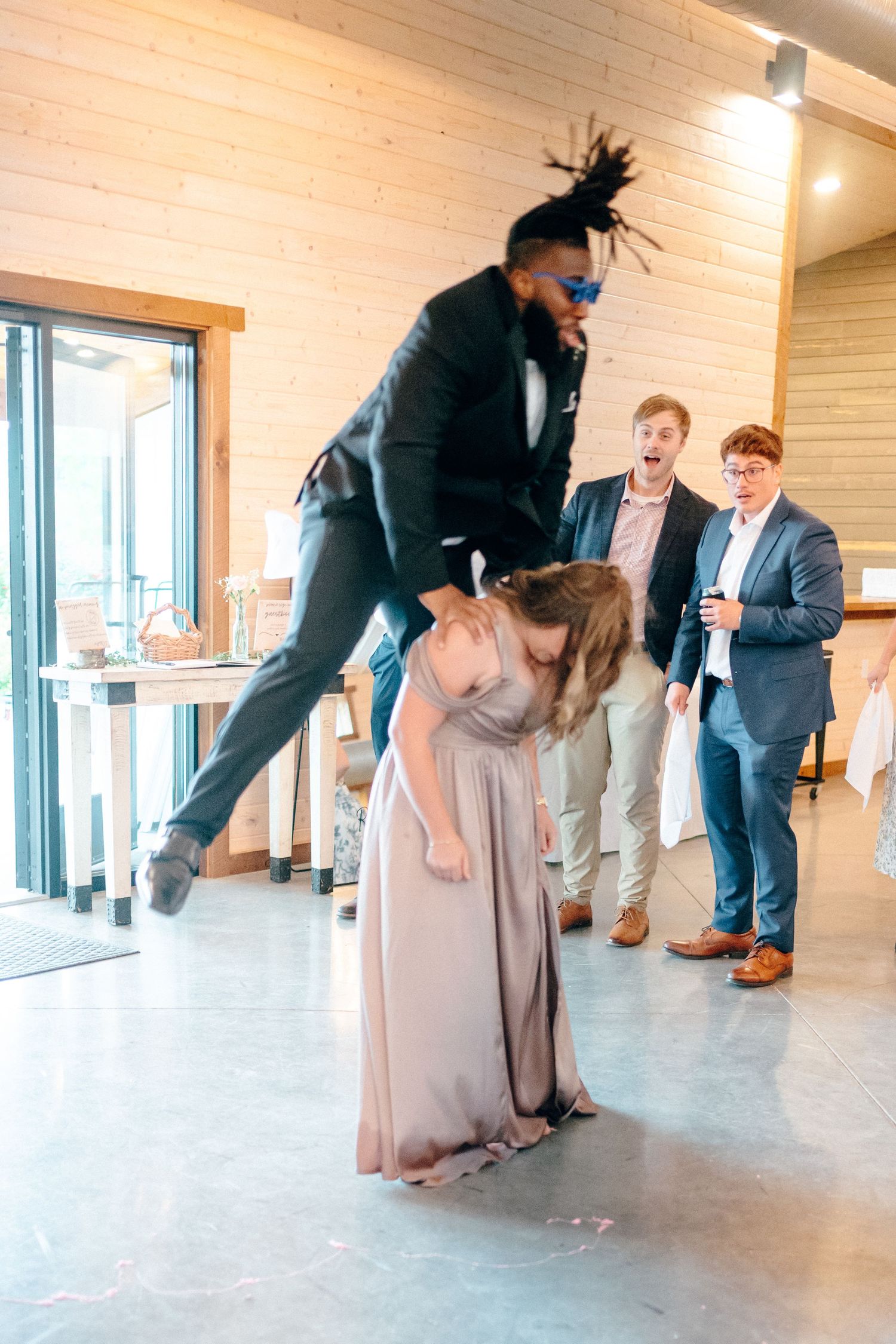 Wedding party members perform energetic dance moves in modern venue with wood paneled walls.