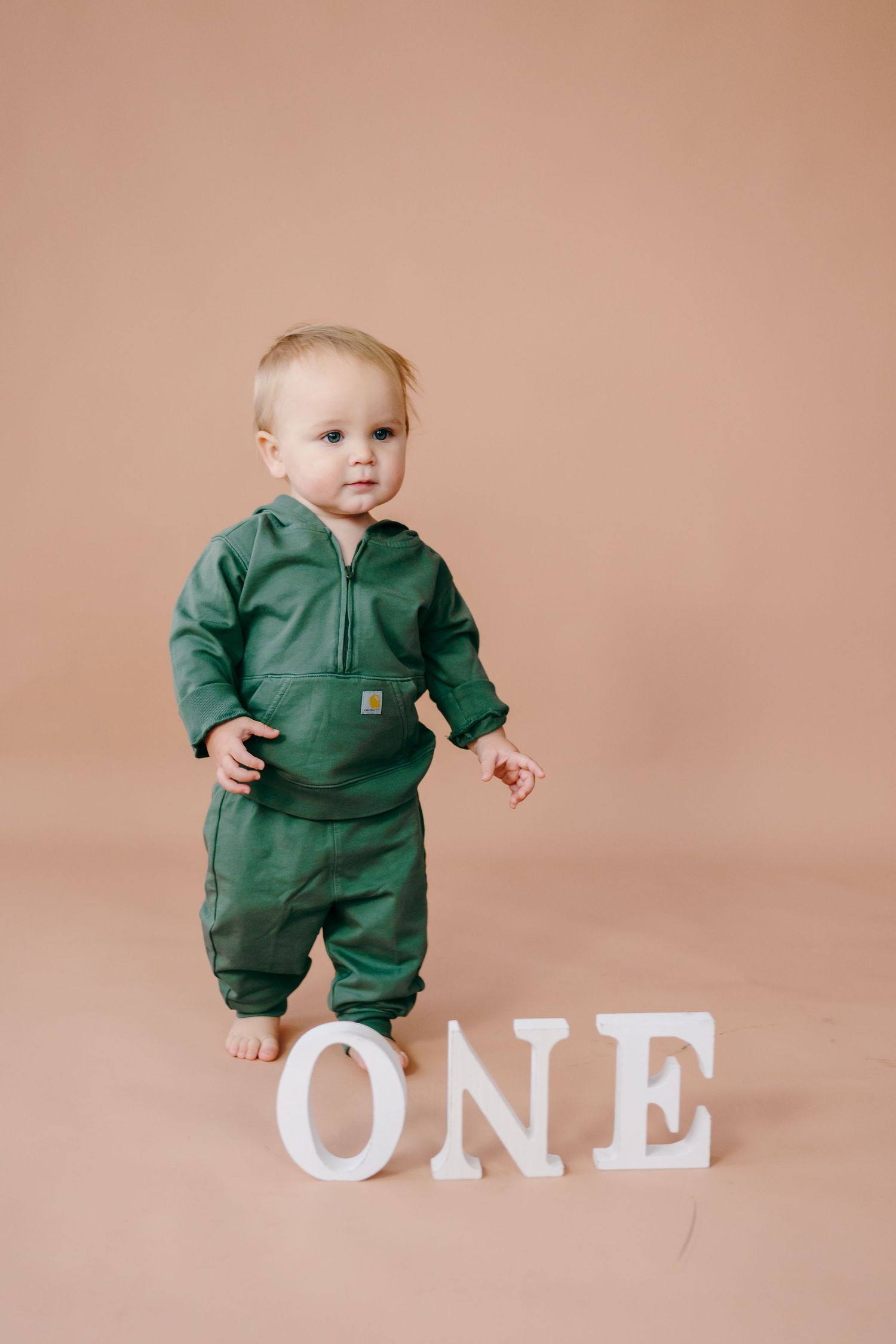 Baby in green Carhartt jumpsuit poses with white ONE letters against beige background.