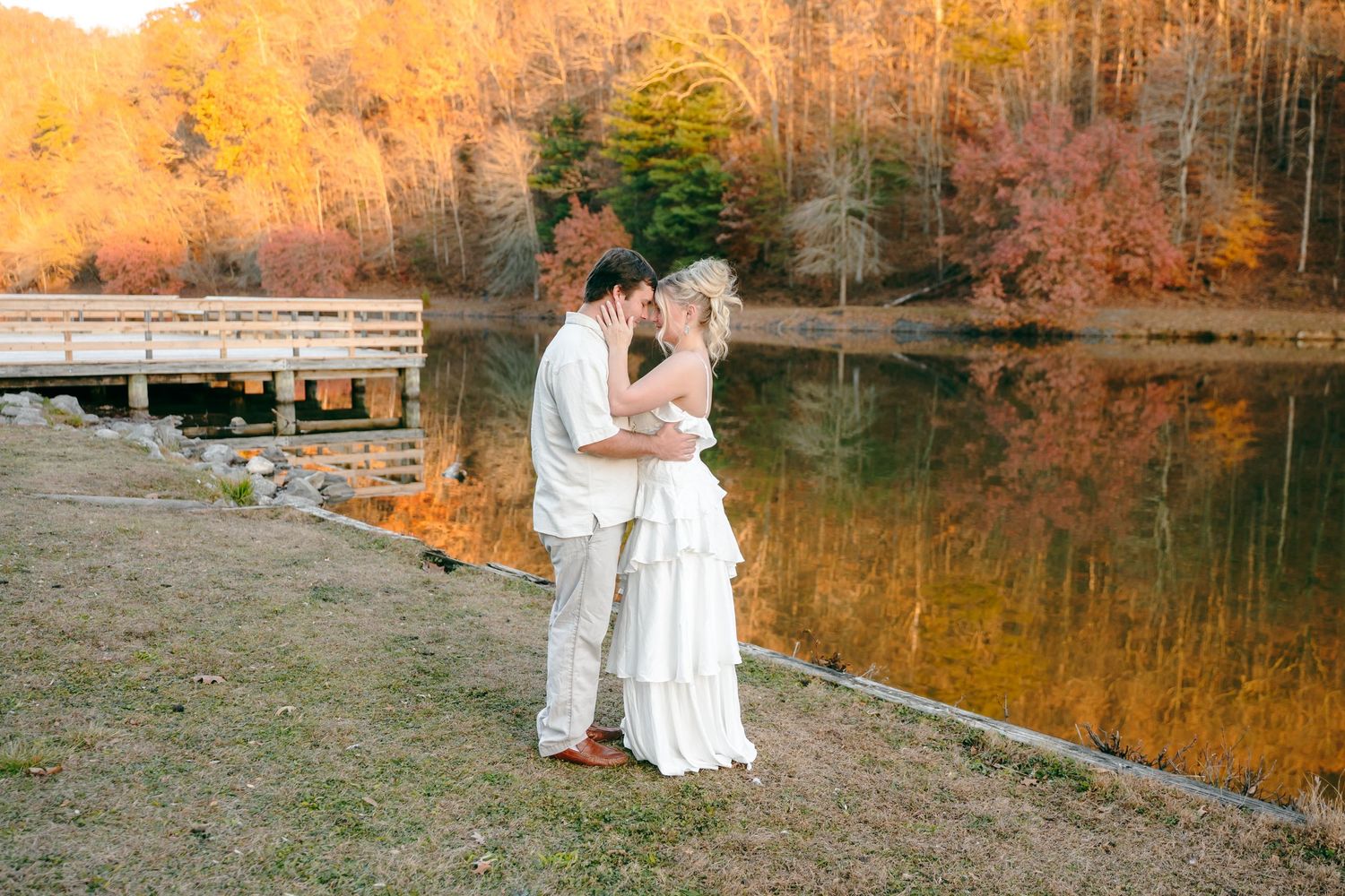 A romantic couple in white outfits embraces by a lake with vibrant fall foliage reflecting in the water.