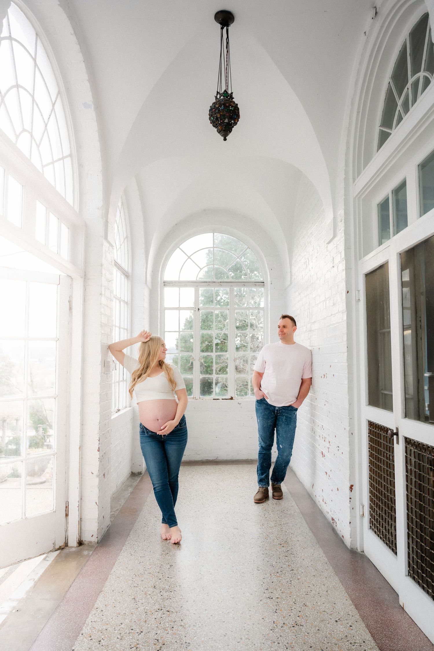 Couple poses in bright white hallway with arched windows and architectural details.