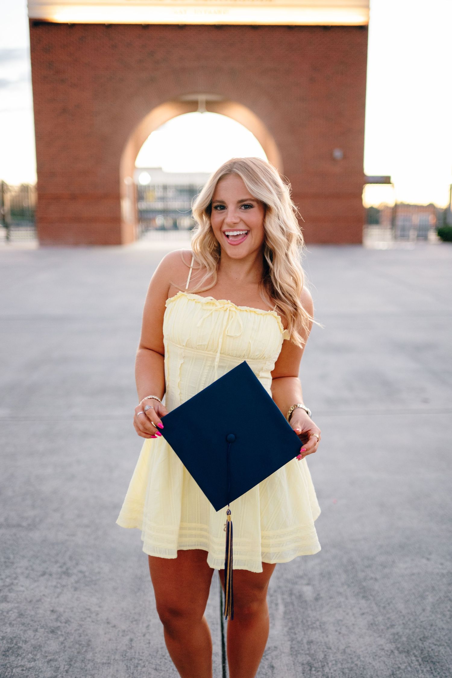 Graduate in yellow dress holding diploma and posing for celebratory photos on urban campus.