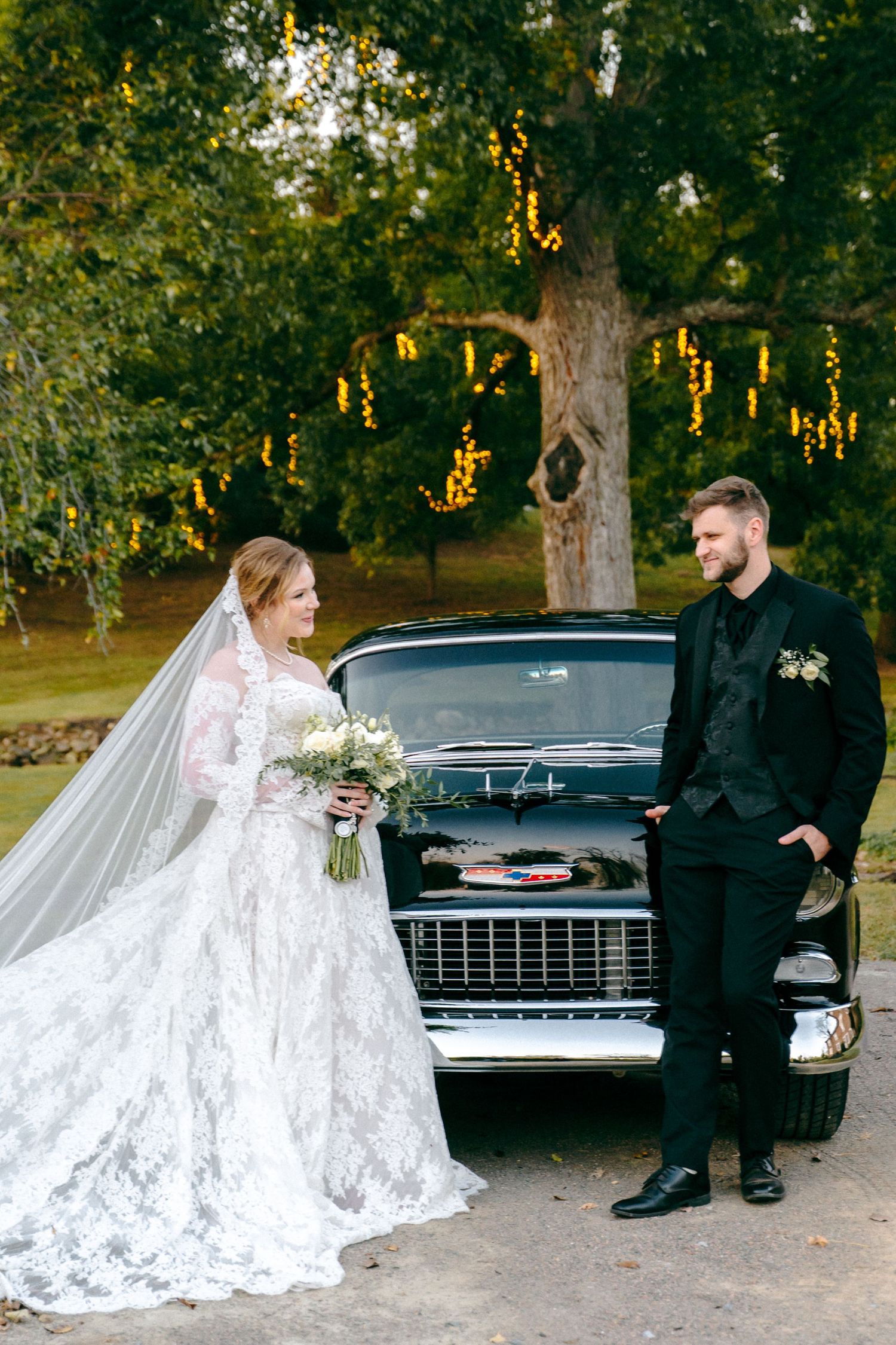 Wedding portraits in a park at sunset with a vintage black car and elegant lace gown.