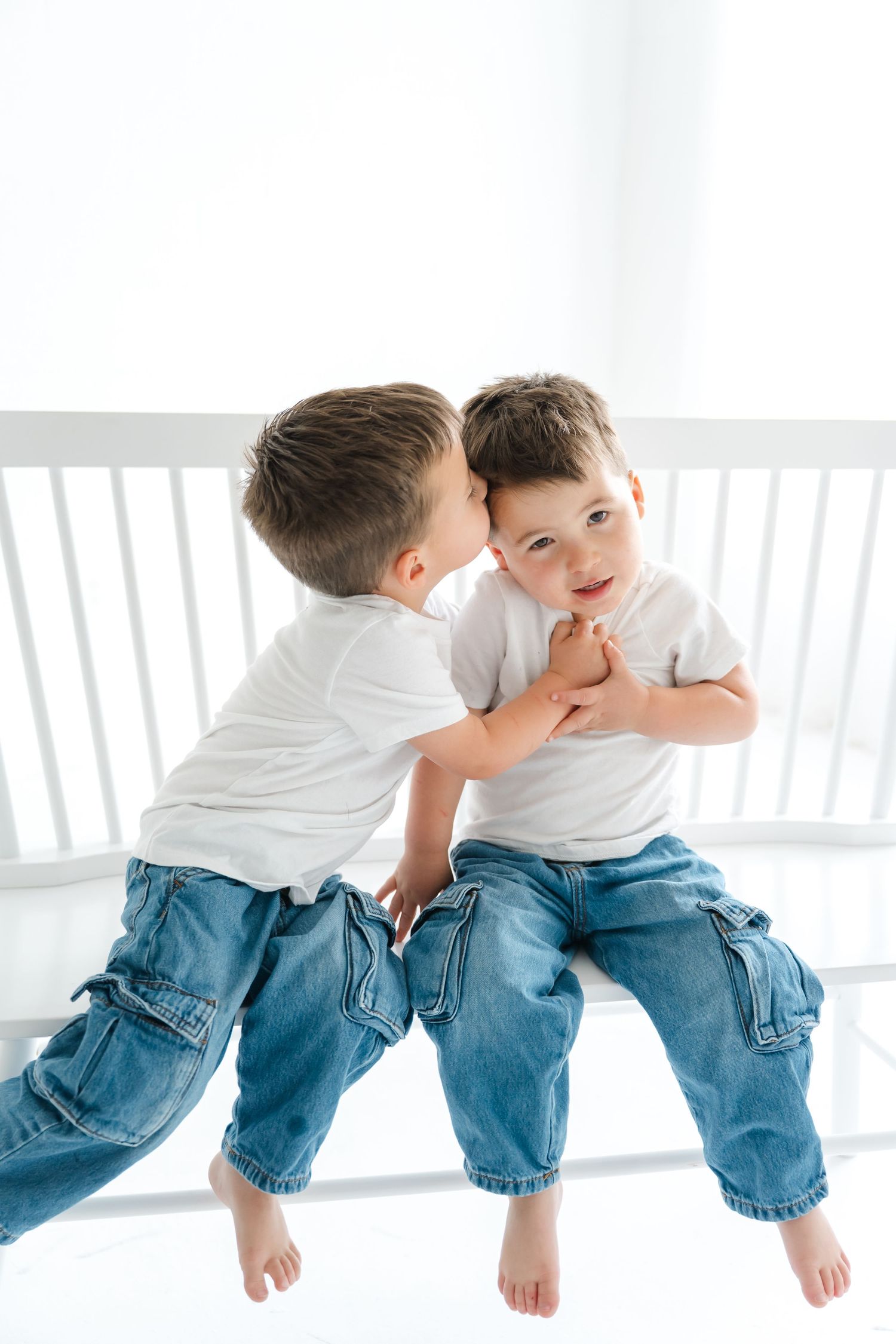 Two young brothers in matching white shirts and jeans sit together, one whispering in the other's ear.