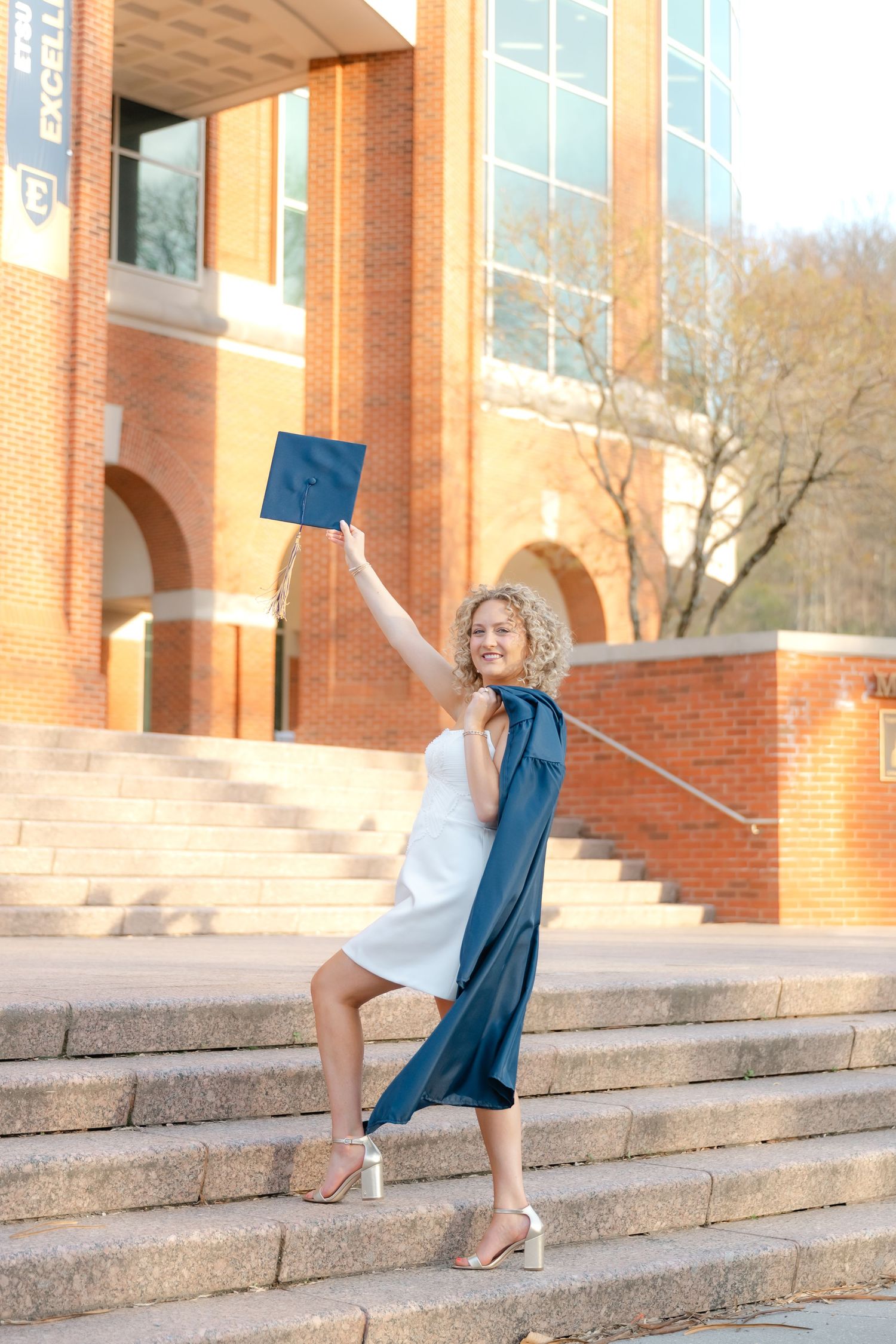 Smiling female graduate in white dress and blue sash raises cap on university steps in golden hour light.