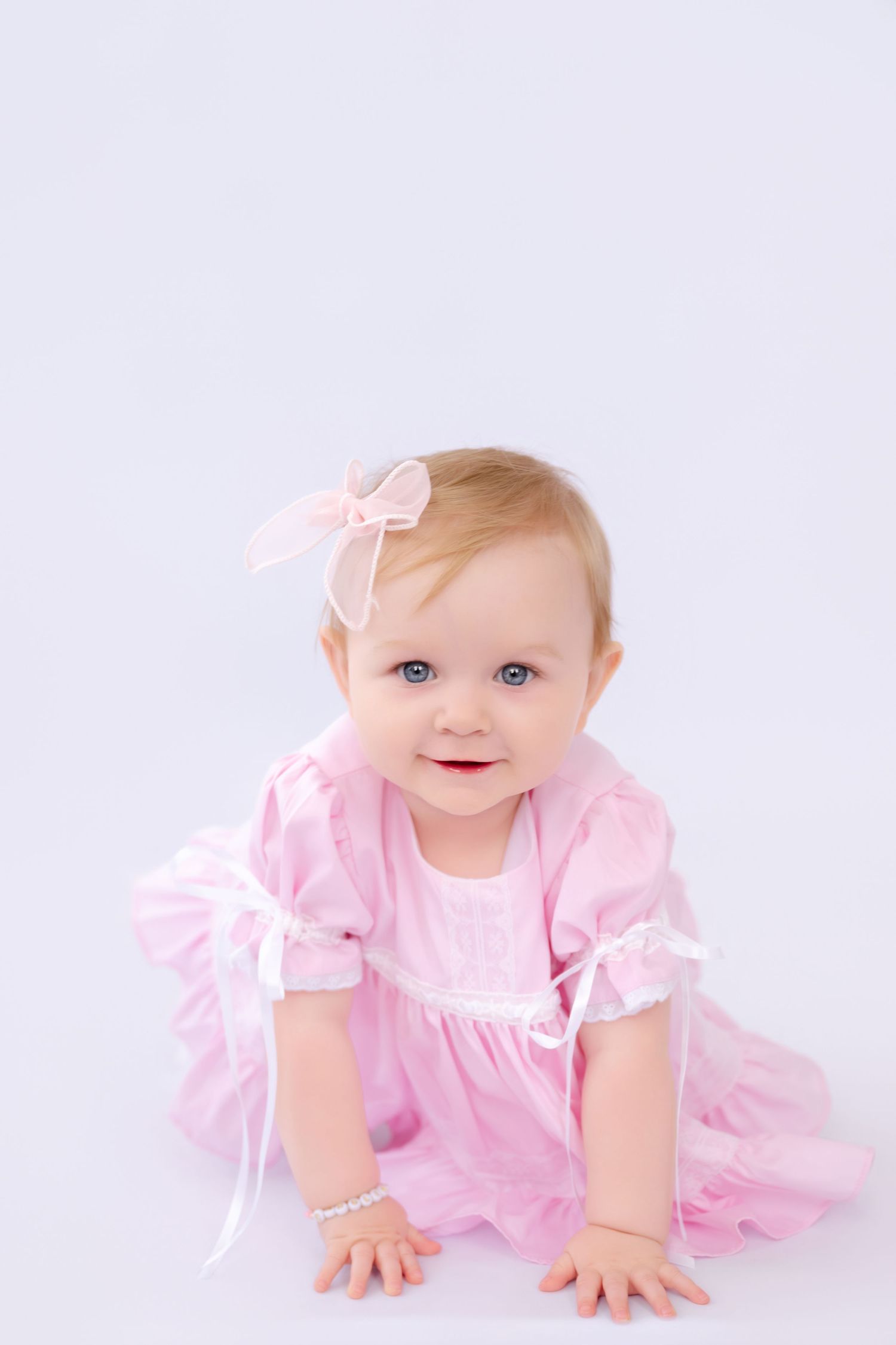 Adorable baby girl in pink dress and bow headband with blue eyes crawling on white background.