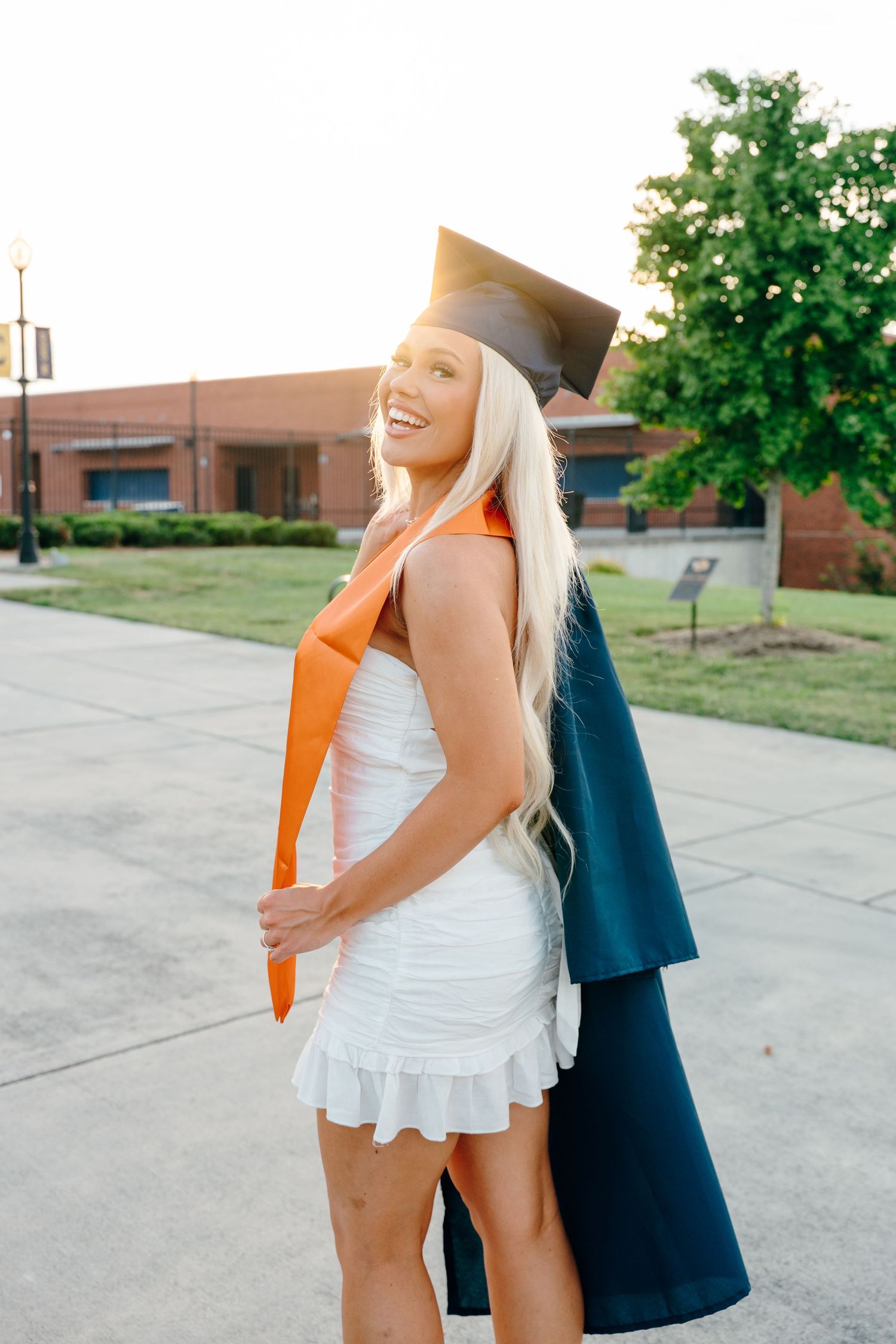 Graduate celebrates with orange stole and white dress in outdoor campus setting.