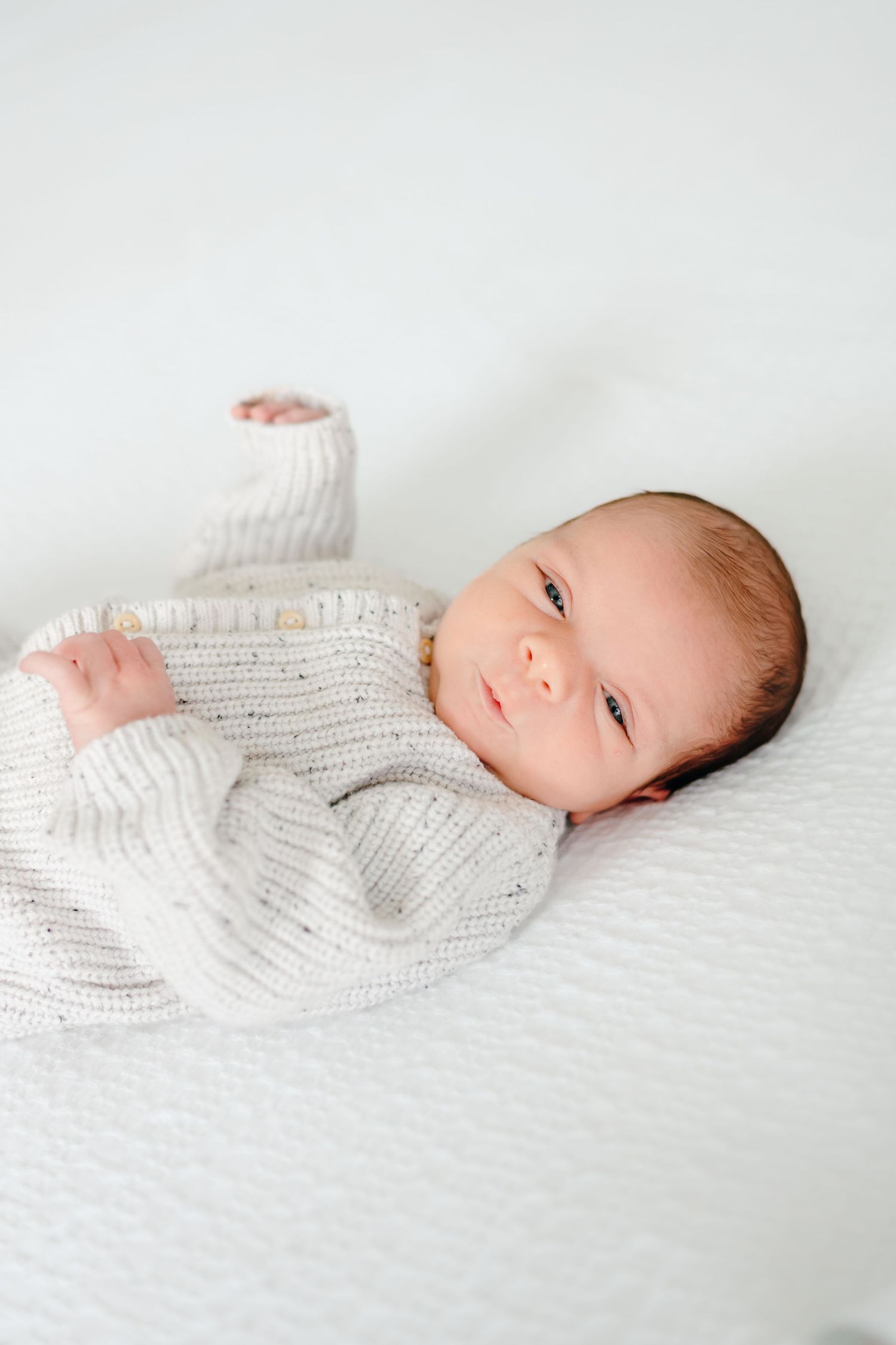 Baby in white sweater lies on textured white bedding looking up at camera.