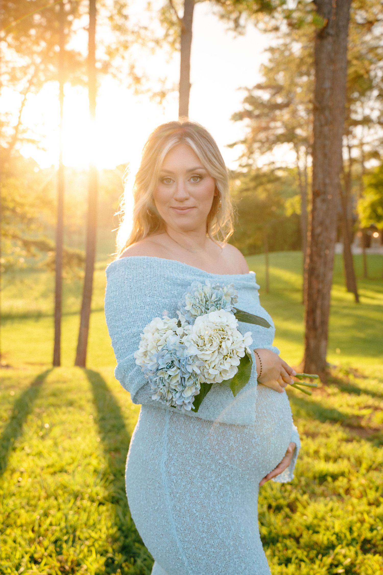 Maternity photoshoot in a sunlit forest featuring a light blue dress and white floral bouquet.
