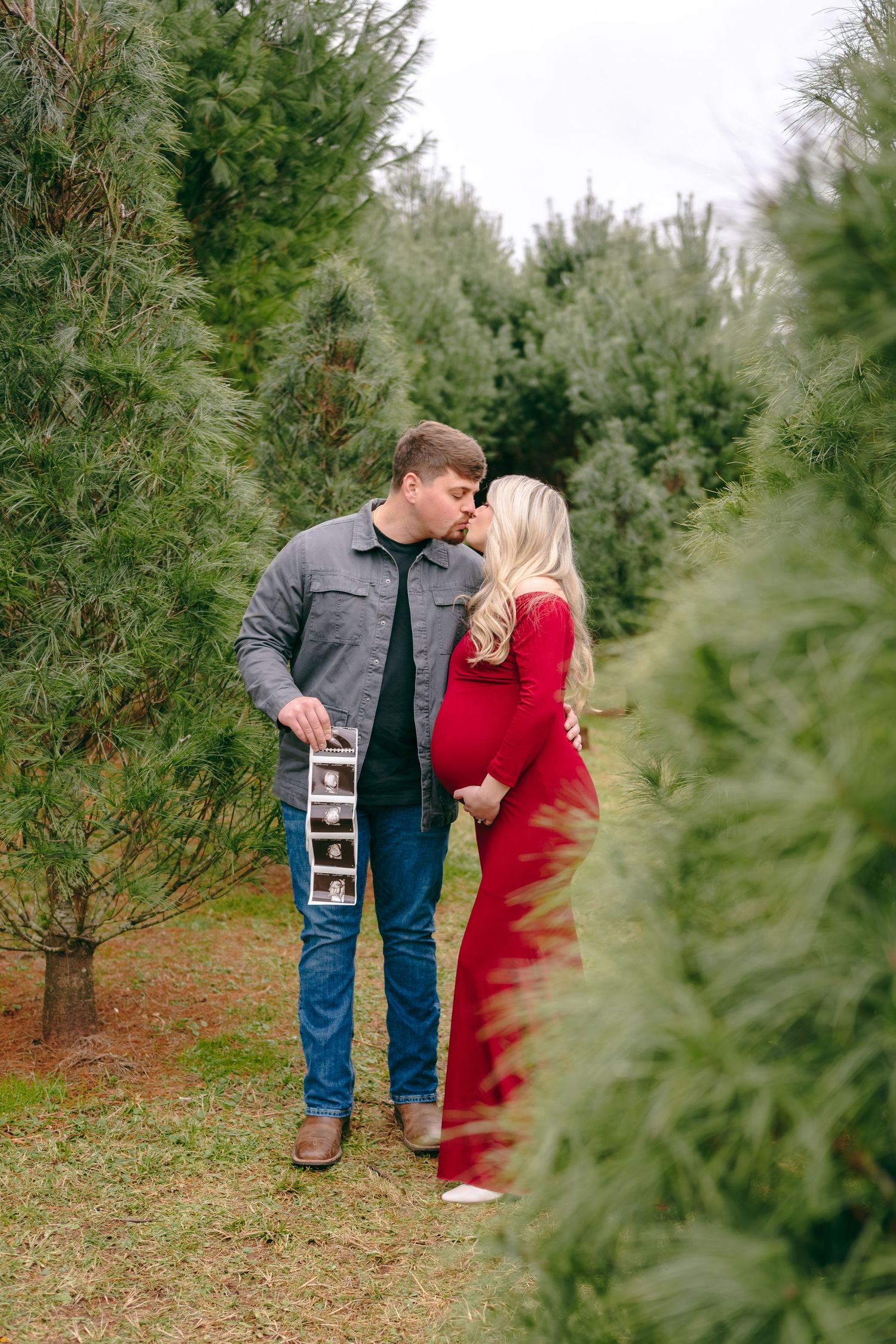 Couple holding ultrasound strip poses among evergreen trees at Christmas tree farm.