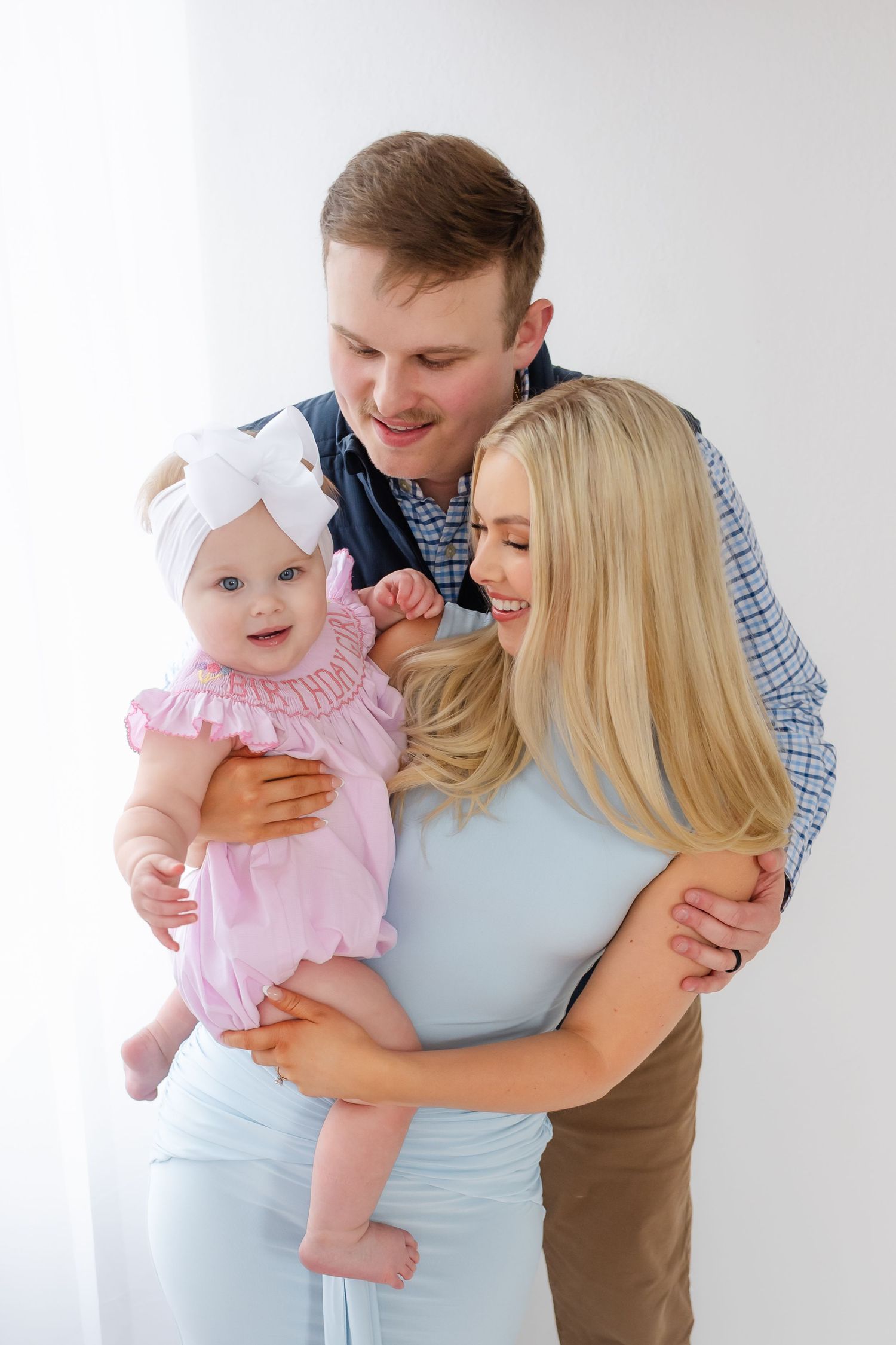 A couple in casual attire share tender moments with a baby wearing pink ruffles against a white background.