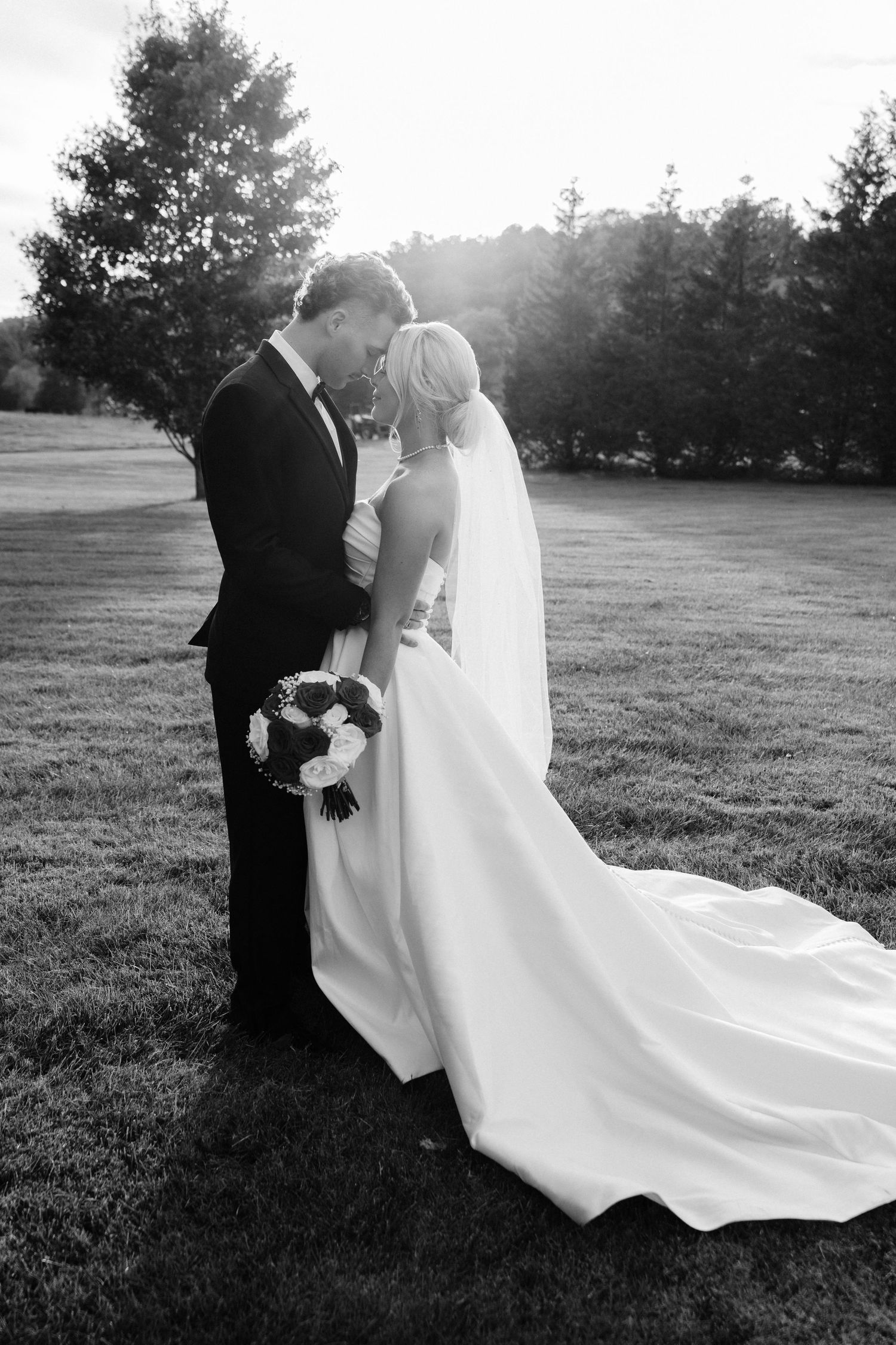Black and white portrait of newlyweds walking through a sunset-lit field.