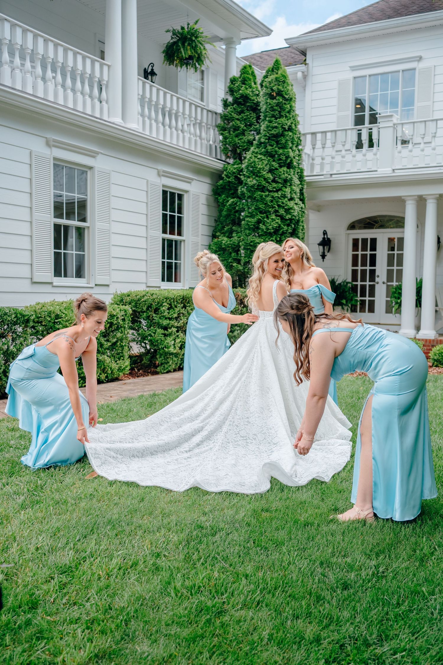 Bridesmaids in light blue dresses admire bride's wedding gown train on mansion lawn.