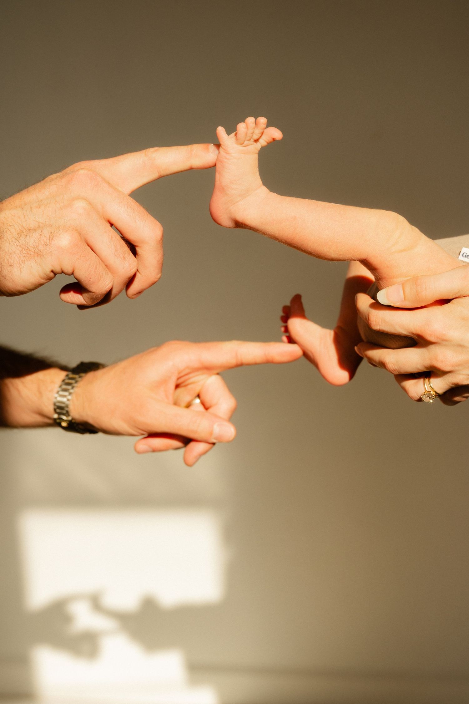 Two parents point fingers at their newborn baby's tiny feet in soft natural light.