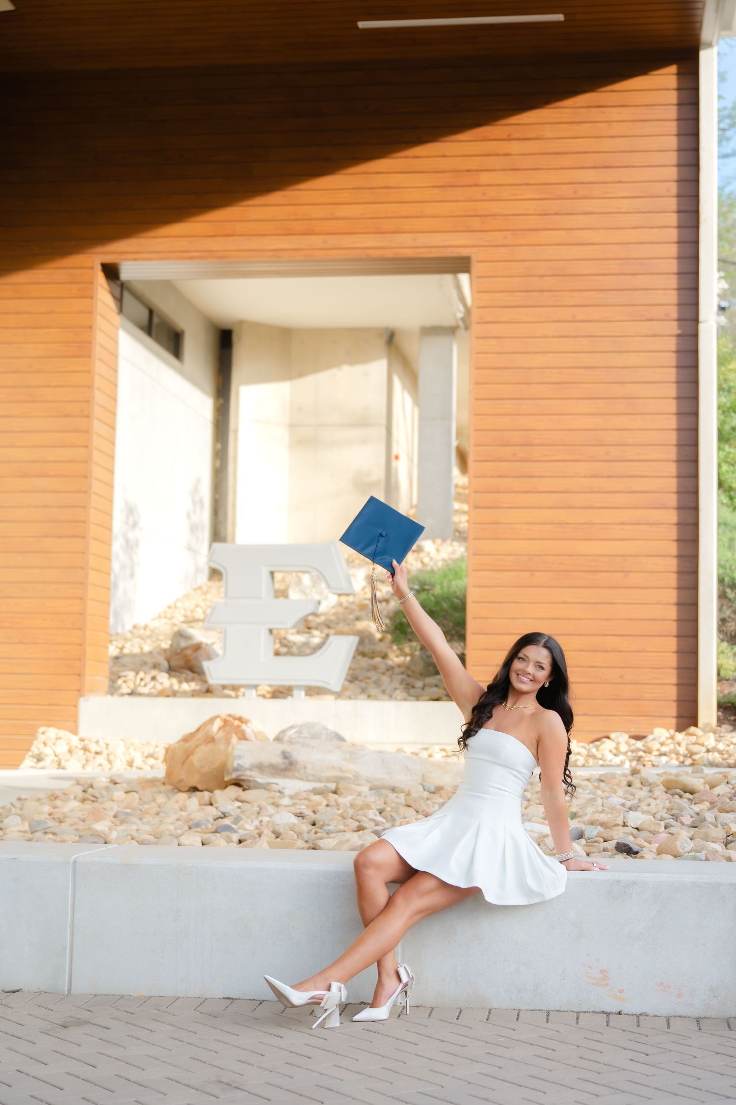 Young woman in white dress holding blue diploma aloft, celebrating graduation while seated outside brick university building.
