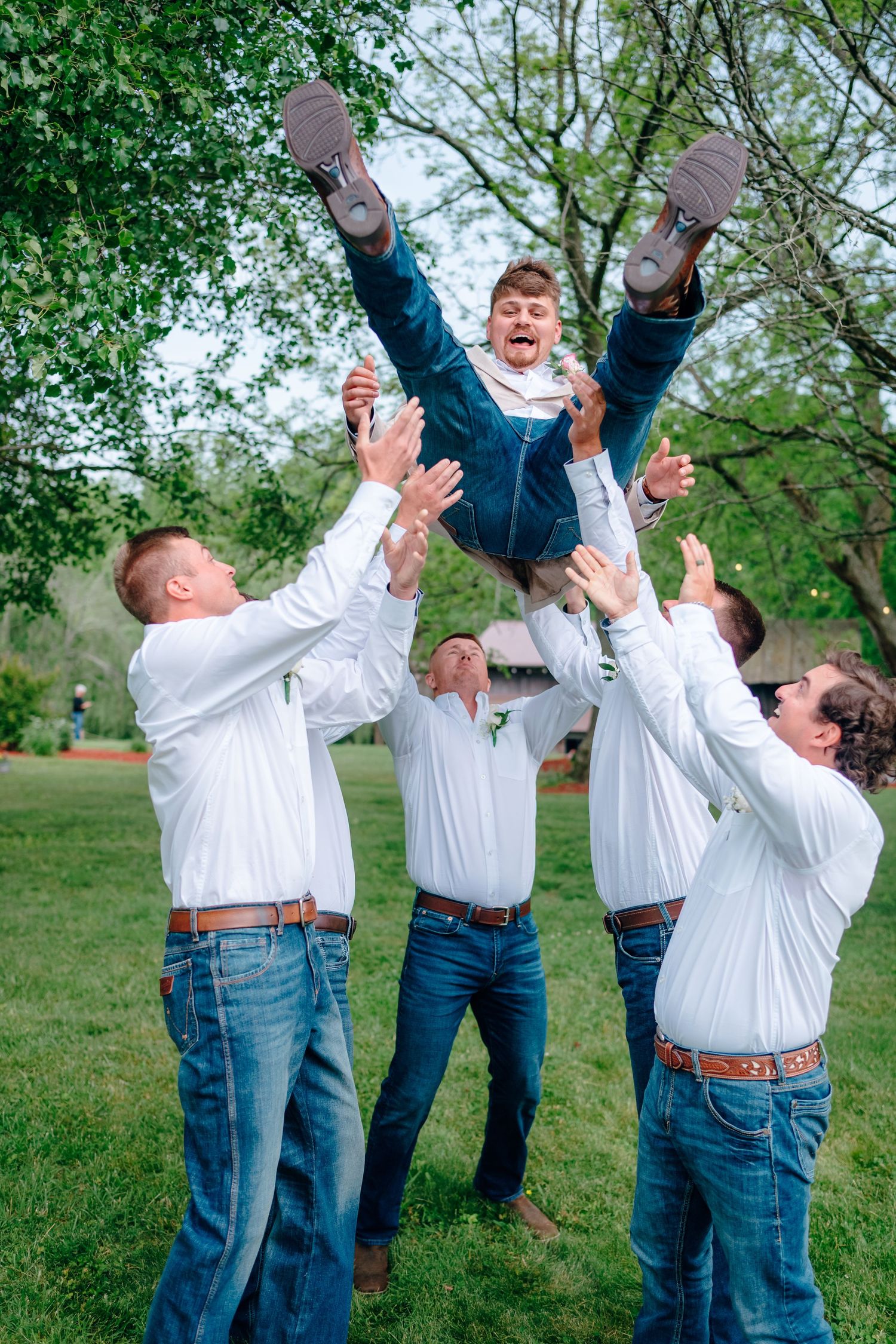 Groomsmen in white shirts and jeans playfully tossing someone in the air outdoors.